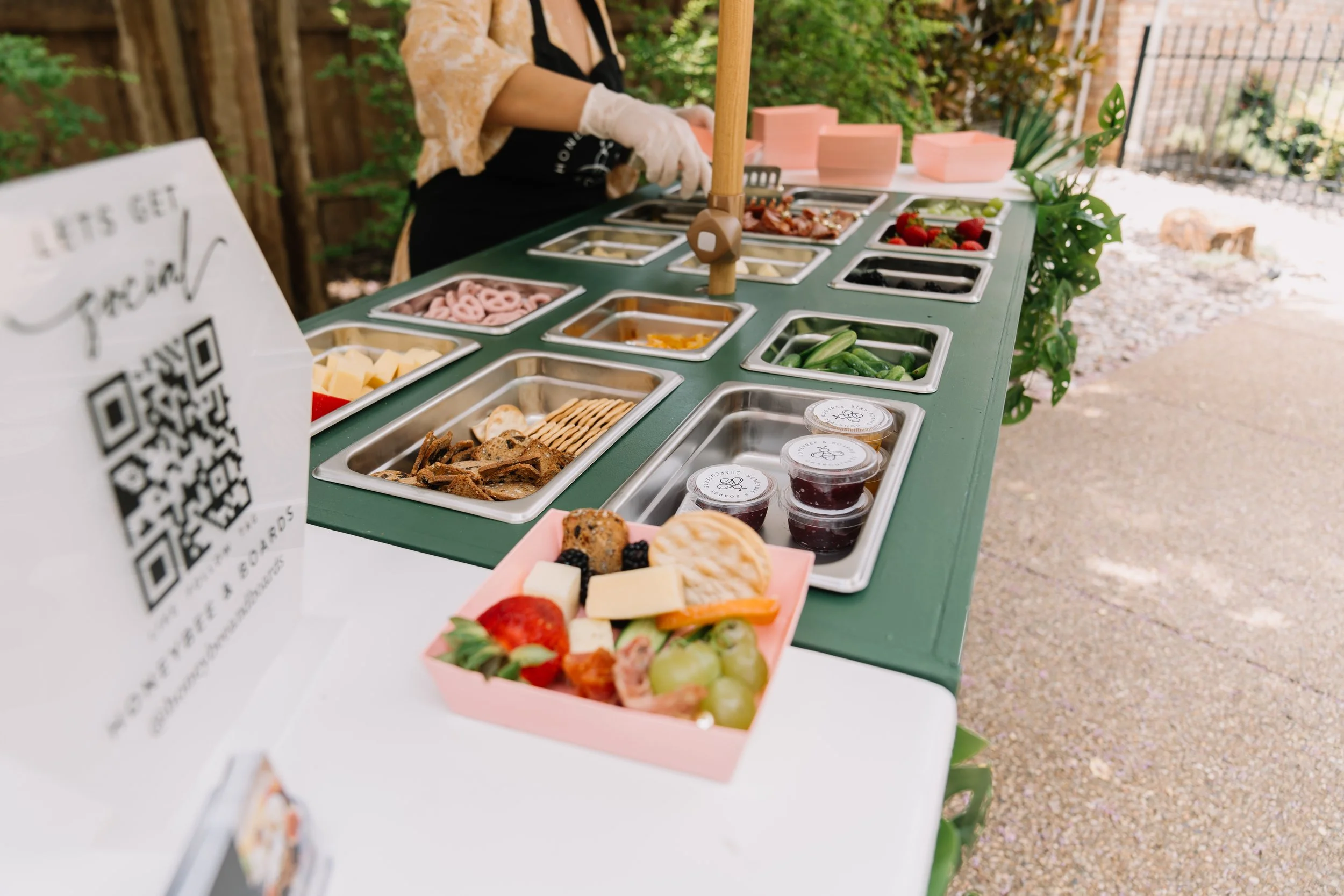 Outdoor breakfast or brunch station with various food containers, fresh fruit, and toppings on a green table, and a person preparing food in the background.