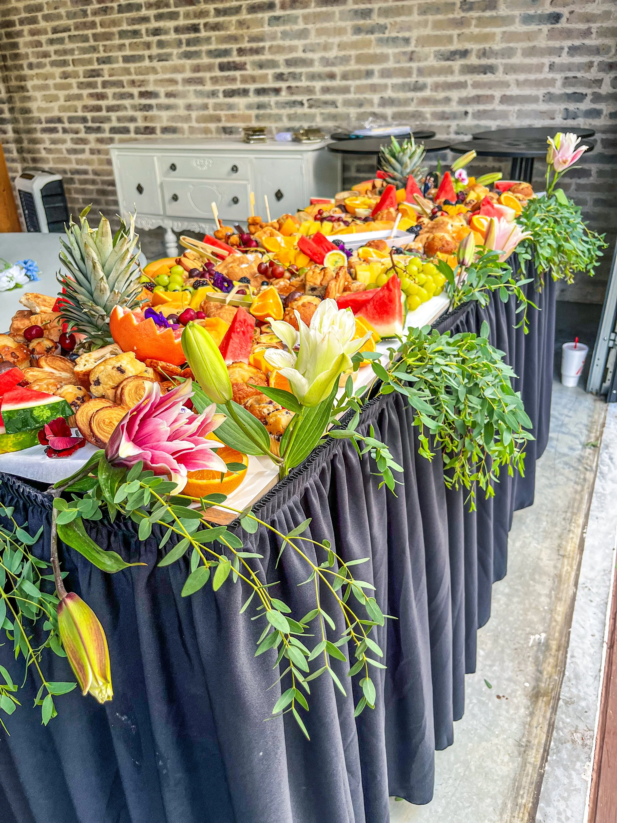 A long table decorated with flowers and topped with an assortment of fruits, pastries, and watermelons, set against a brick wall background.