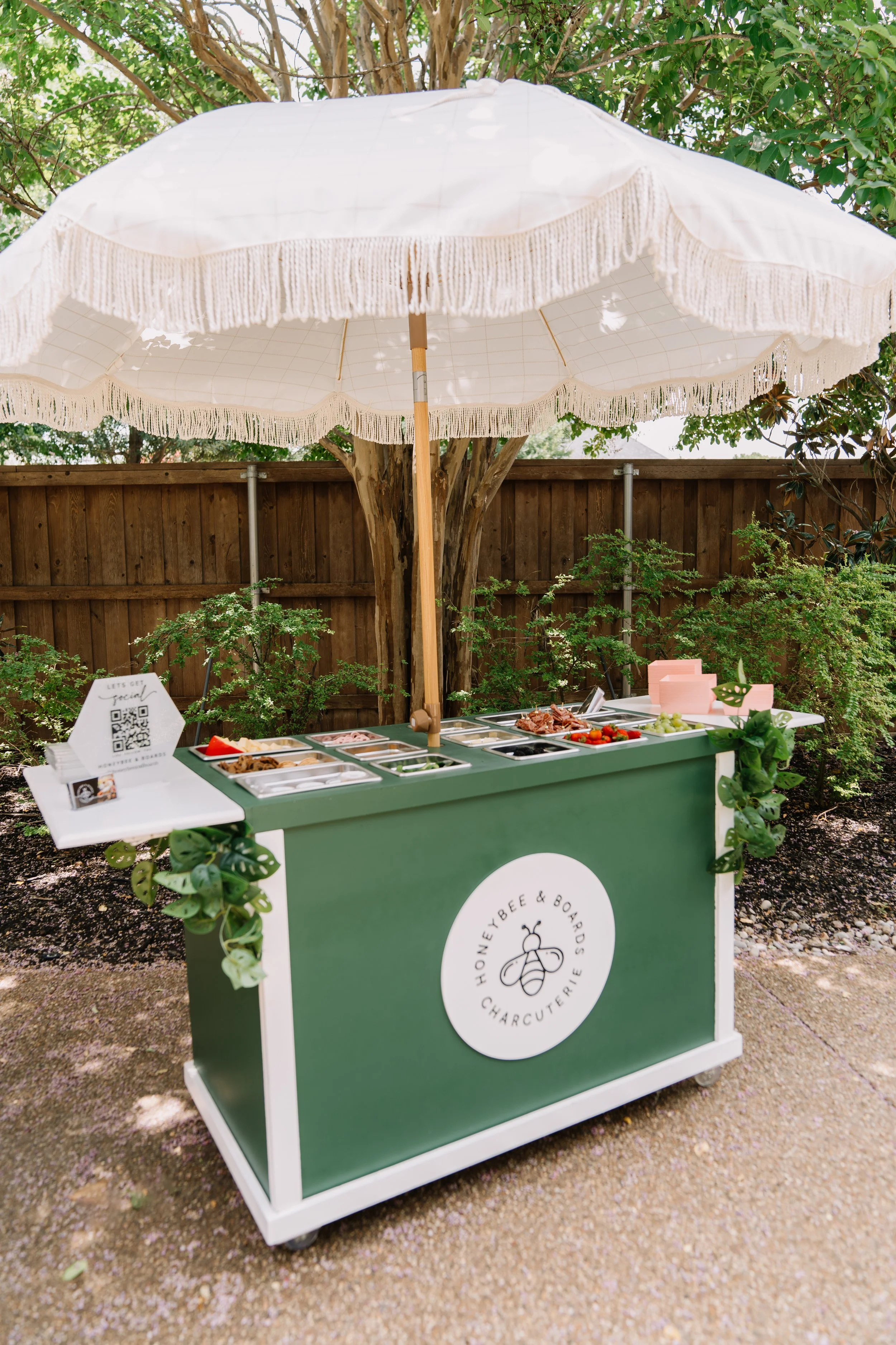A food cart under a large white umbrella with fringes, offering various meats, fruits, and other items in a garden setting with a wooden fence and trees in the background.