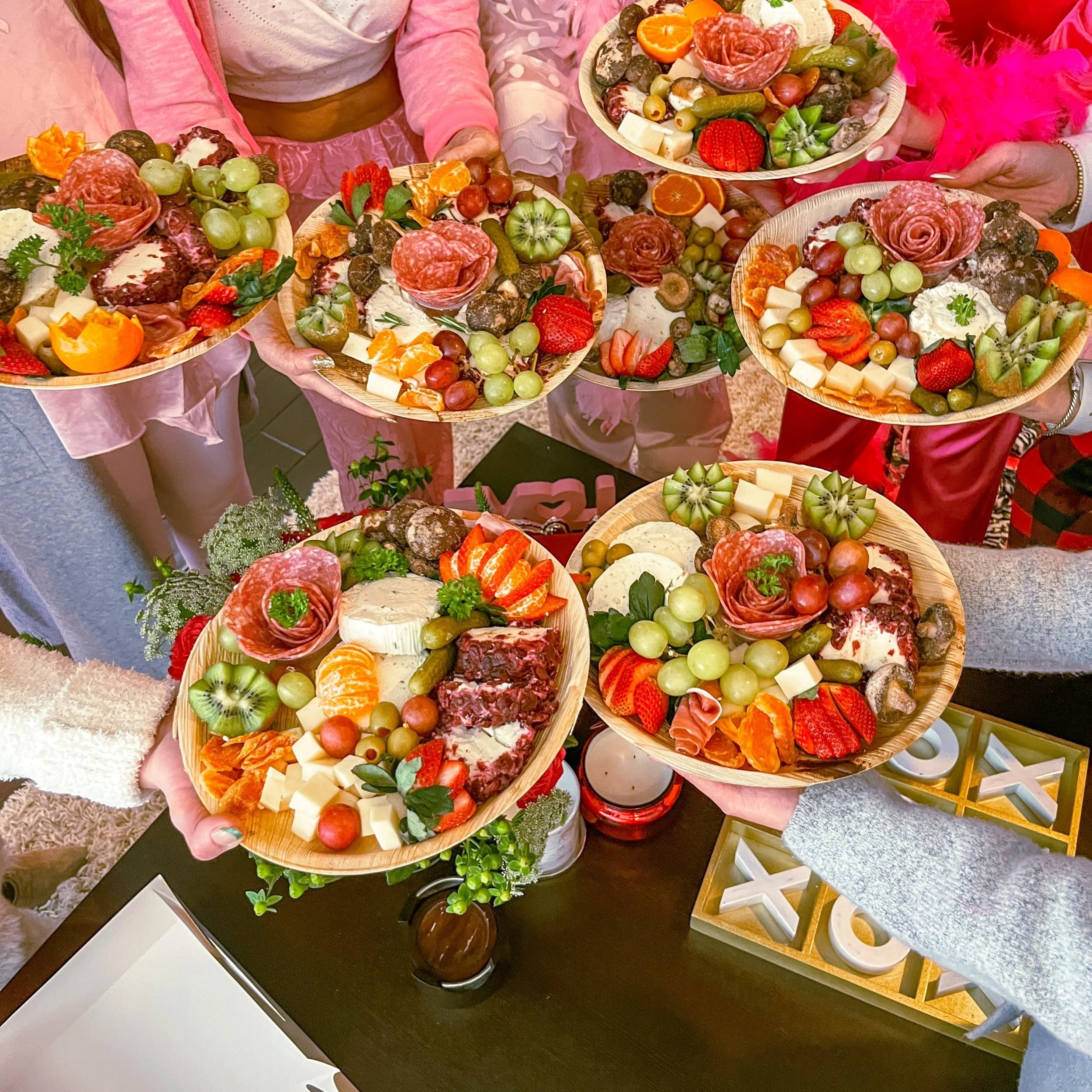 Several people holding wooden plates filled with cheese, grapes, strawberries, kiwis, sliced oranges, cured meats, and vegetables, celebrating together.