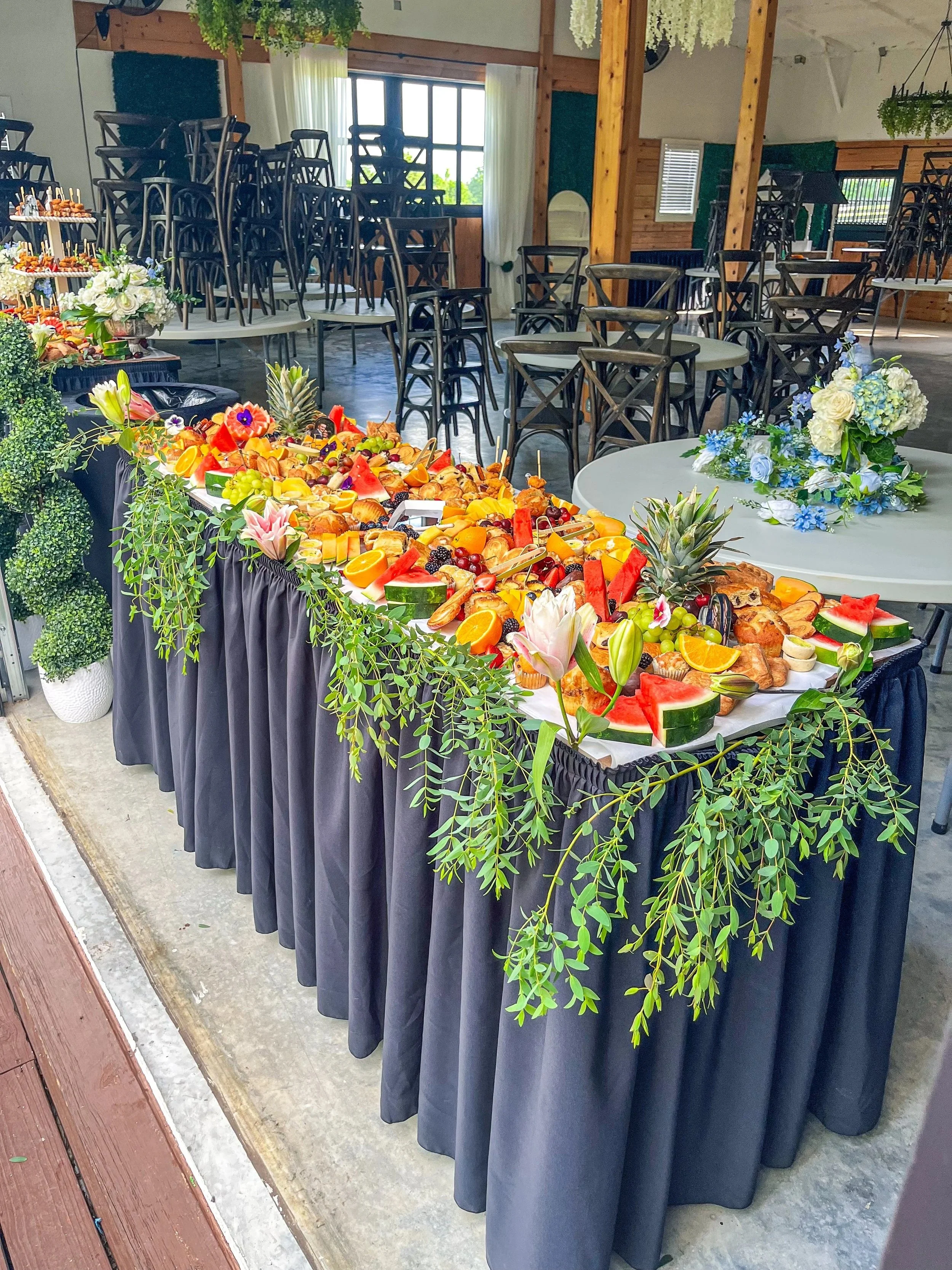 A buffet table decorated with fruits, vegetables, and flowers at an indoor event space. The table has a black skirt and is adorned with green leafy vines and pink lilies. In the background, there are stacked chairs, round tables with flower arrangeme