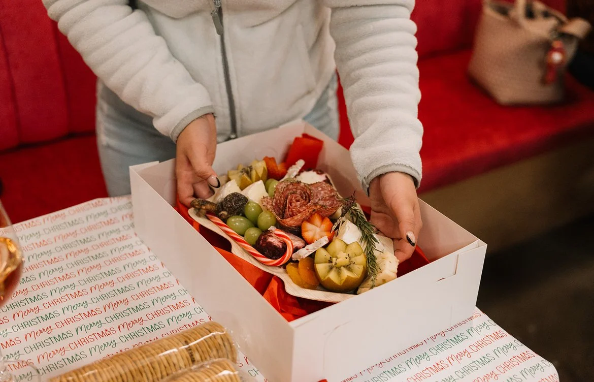 Person holding a white box with assorted Christmas treats and decorations, including grapes, candy canes, strawberries, and rosemary, on a festive table.