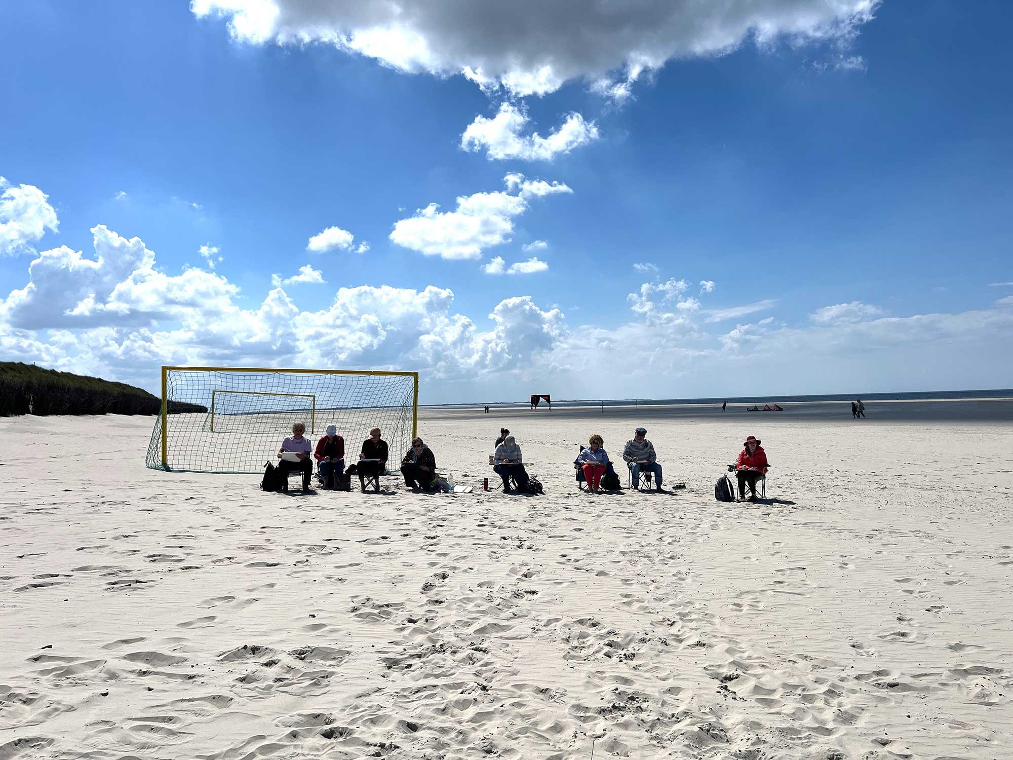 Eine Gruppe von Menschen sitzt mit Stühlen auf einem sandigen Strand vor einem Fußballtor. Im Hintergrund ist das Meer und der Himmel mit Wolken zu sehen.