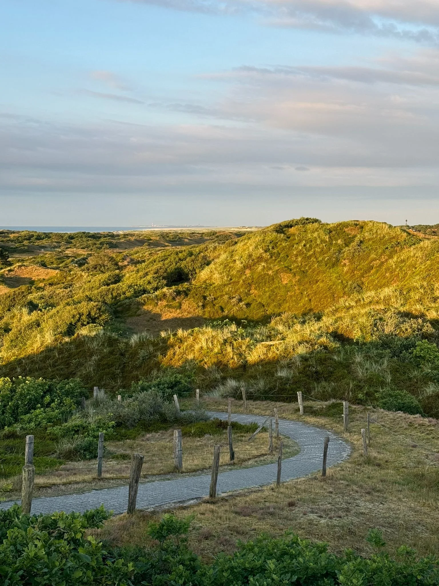 Landschaft mit Hügeln und gepflastertem Weg, umgeben von grüner Vegetation und Gräsern unter blauem Himmel zu sehen. Dezente Abendbeleuchtung erhellt die Szene.