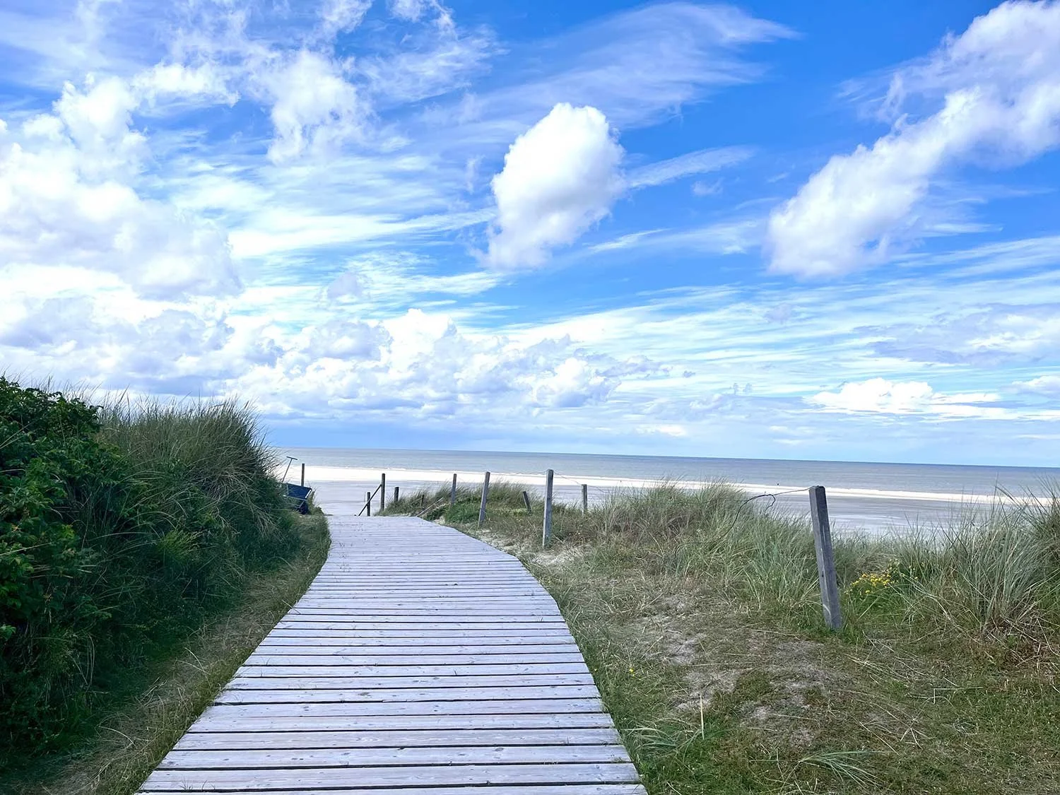 Strandweg durch Dünen, führt zum Meer, blauer Himmel mit Wolken.