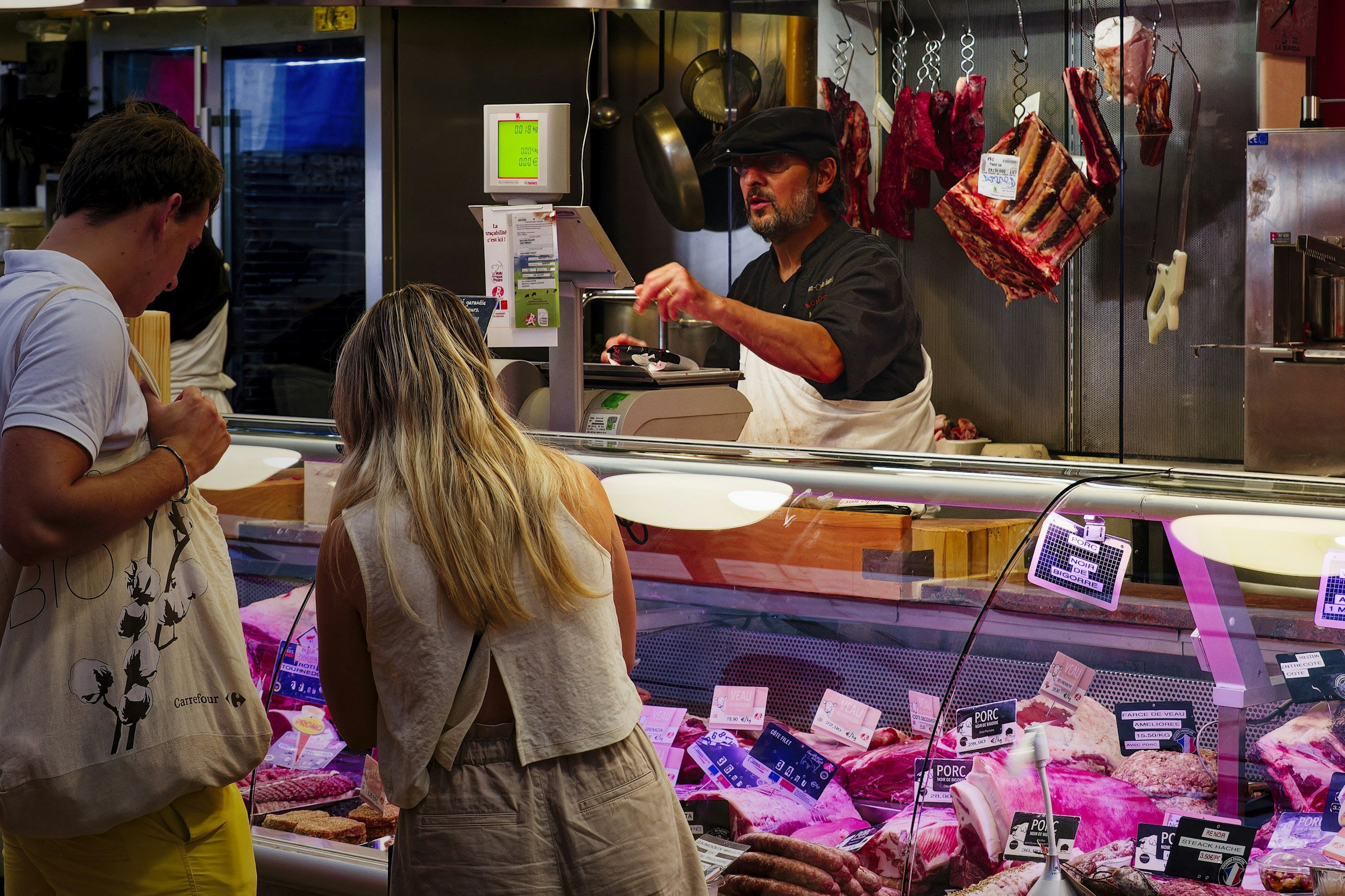 People shopping at a butcher shop with hanging meats and branded labels on the meat cuts.