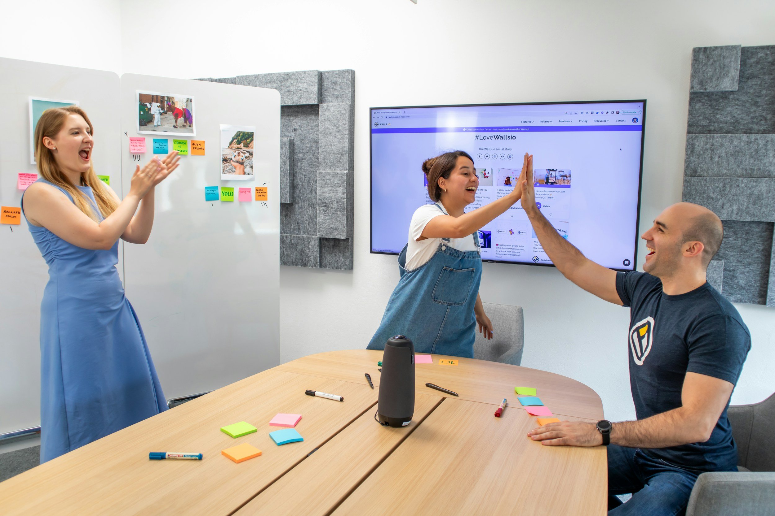 Three people in a meeting room celebrating with high fives. A woman on the left is clapping and smiling, while a woman in the center and a man on the right are giving each other a high five. The table has colorful sticky notes, pens, and a speaker on it. A large screen in the background displays a webpage.