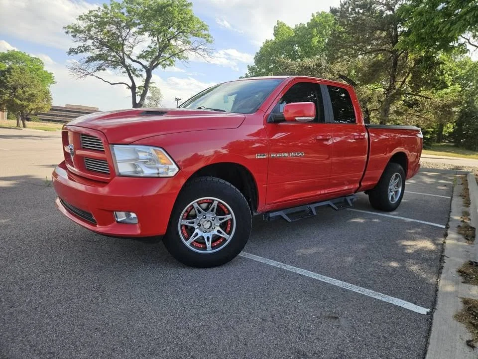A red RAM 1500 pickup truck parked in an outdoor parking lot with trees and a partly cloudy sky in the background.