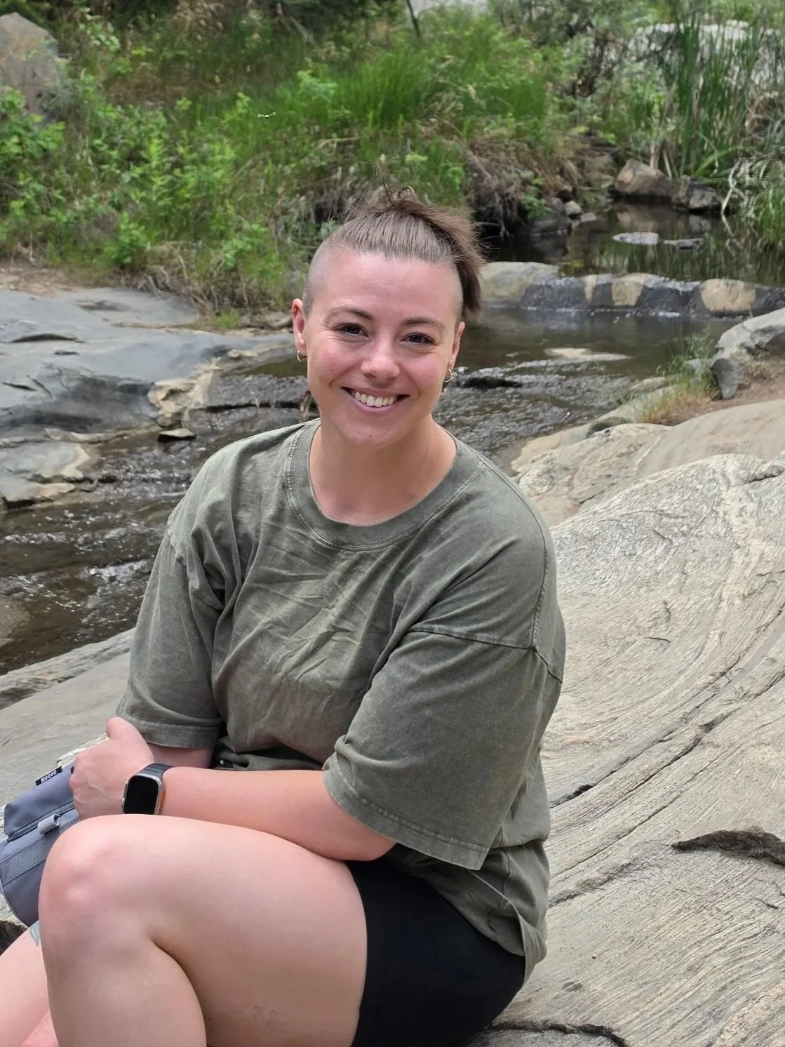 A woman sitting on a large rock next to a small stream with greenery in the background, smiling at the camera.