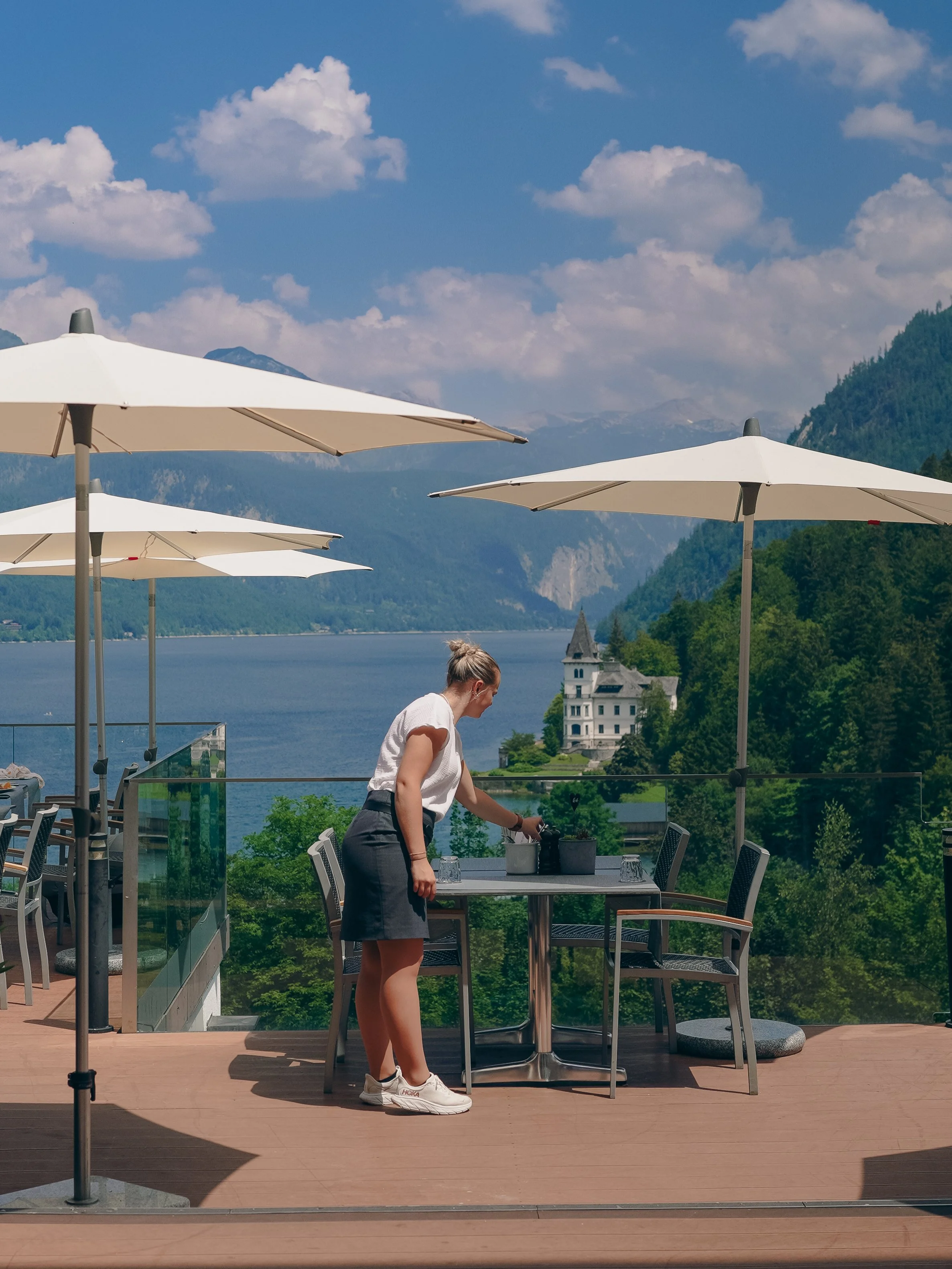 Person setting a table on a patio with umbrellas overlooking a lake and mountains.
