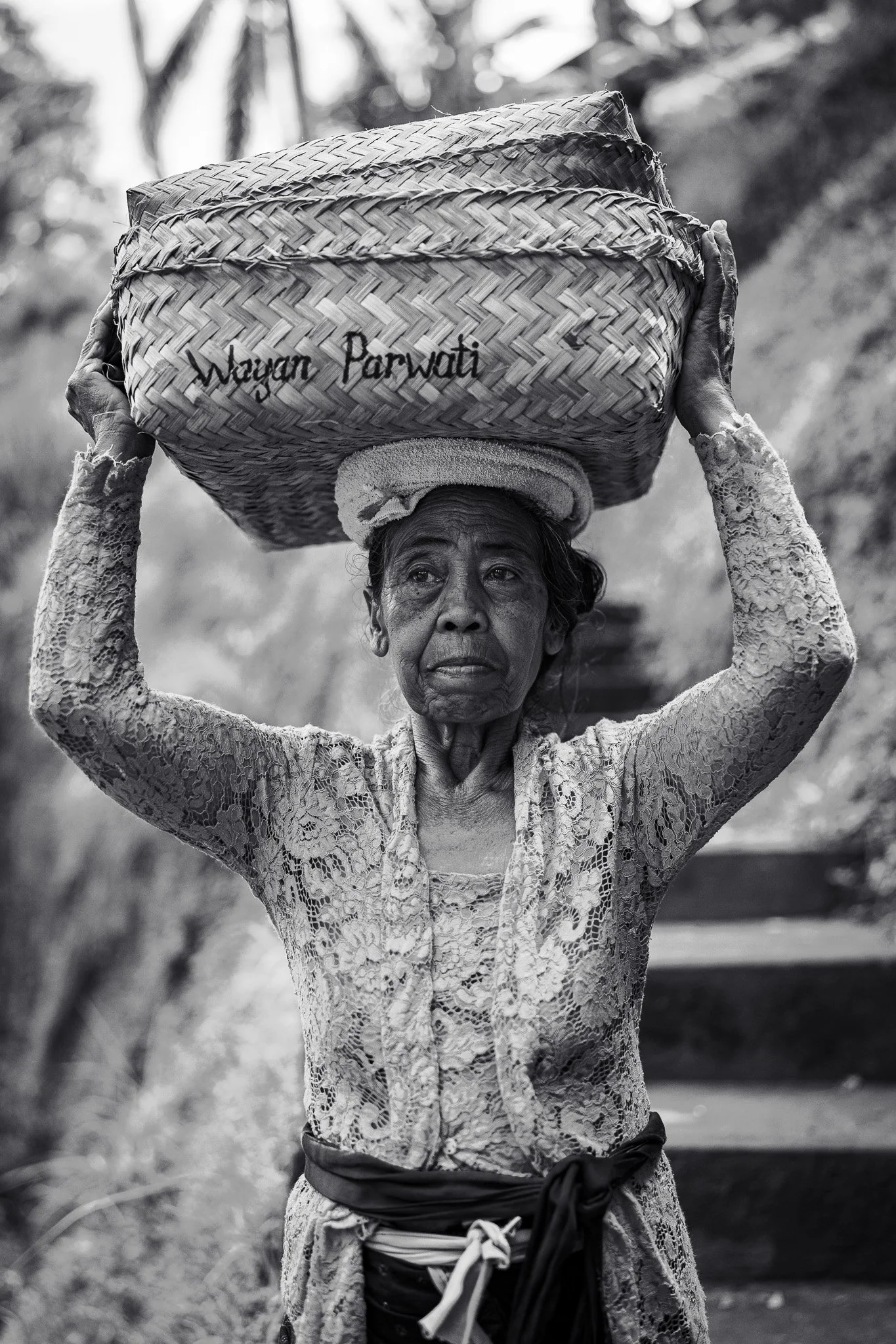 An elderly woman carries a woven basket on her head while standing on a stone path. The basket has text written on it. She wears a traditional lace outfit.