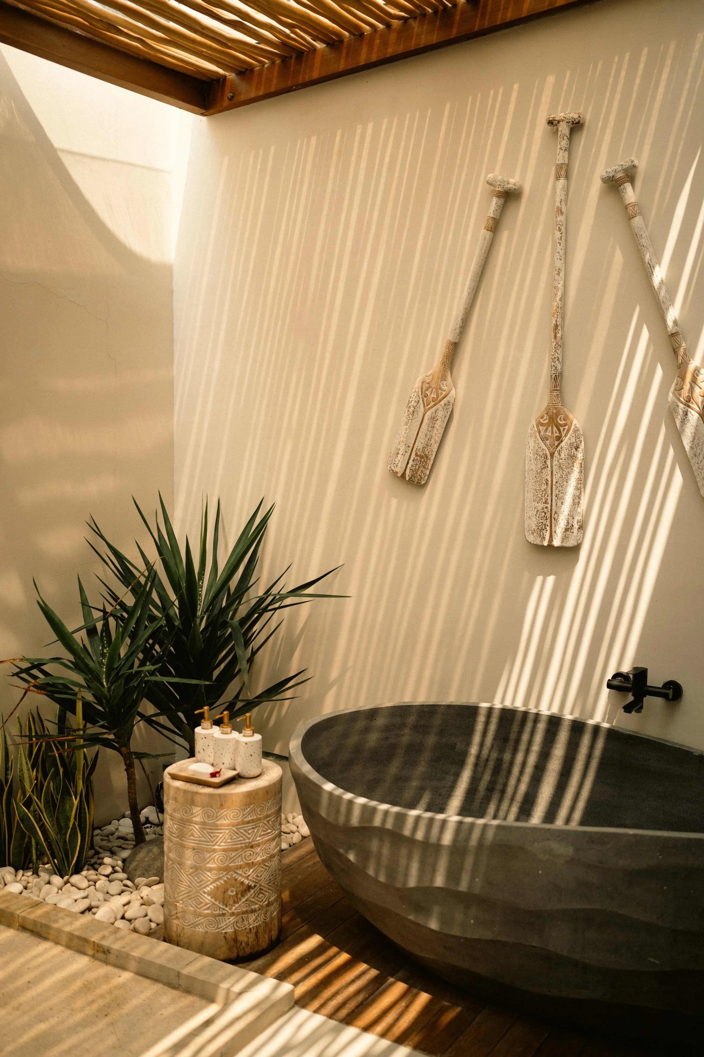 Outdoor bathroom with woven wood ceiling, stone bathtub, decorative oars on wall, potted plants, and soap dispensers on carved stone stool.