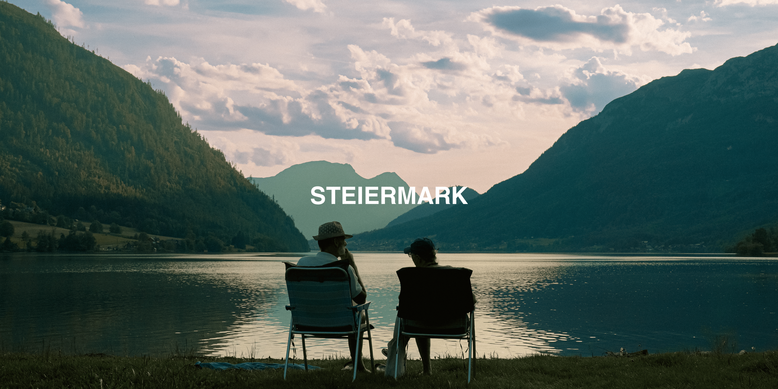 Two people sitting in chairs by a lake with mountains in the background, labeled "Steiermark."