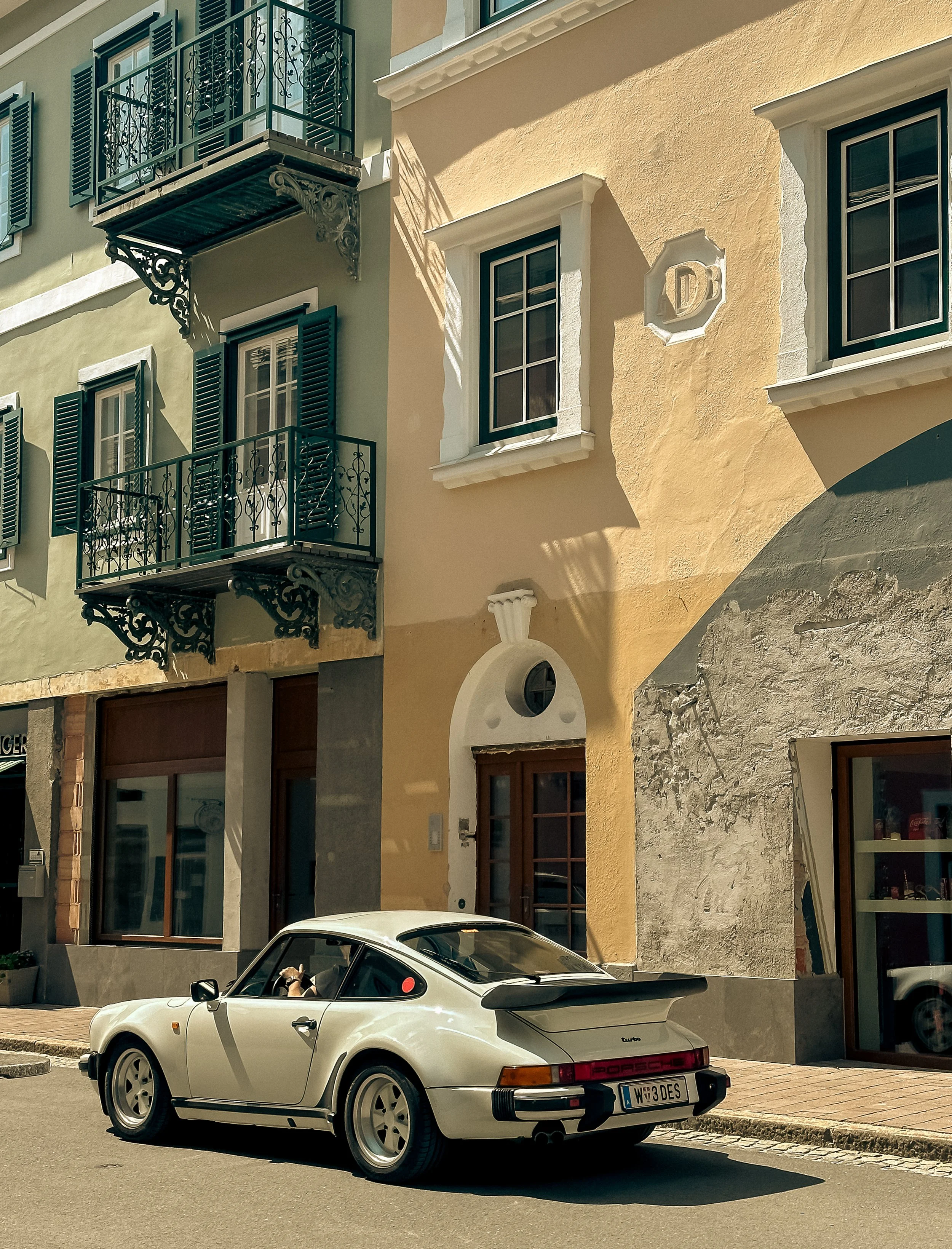 Vintage white Porsche parked on a street with European-style buildings featuring balconies and shutters.