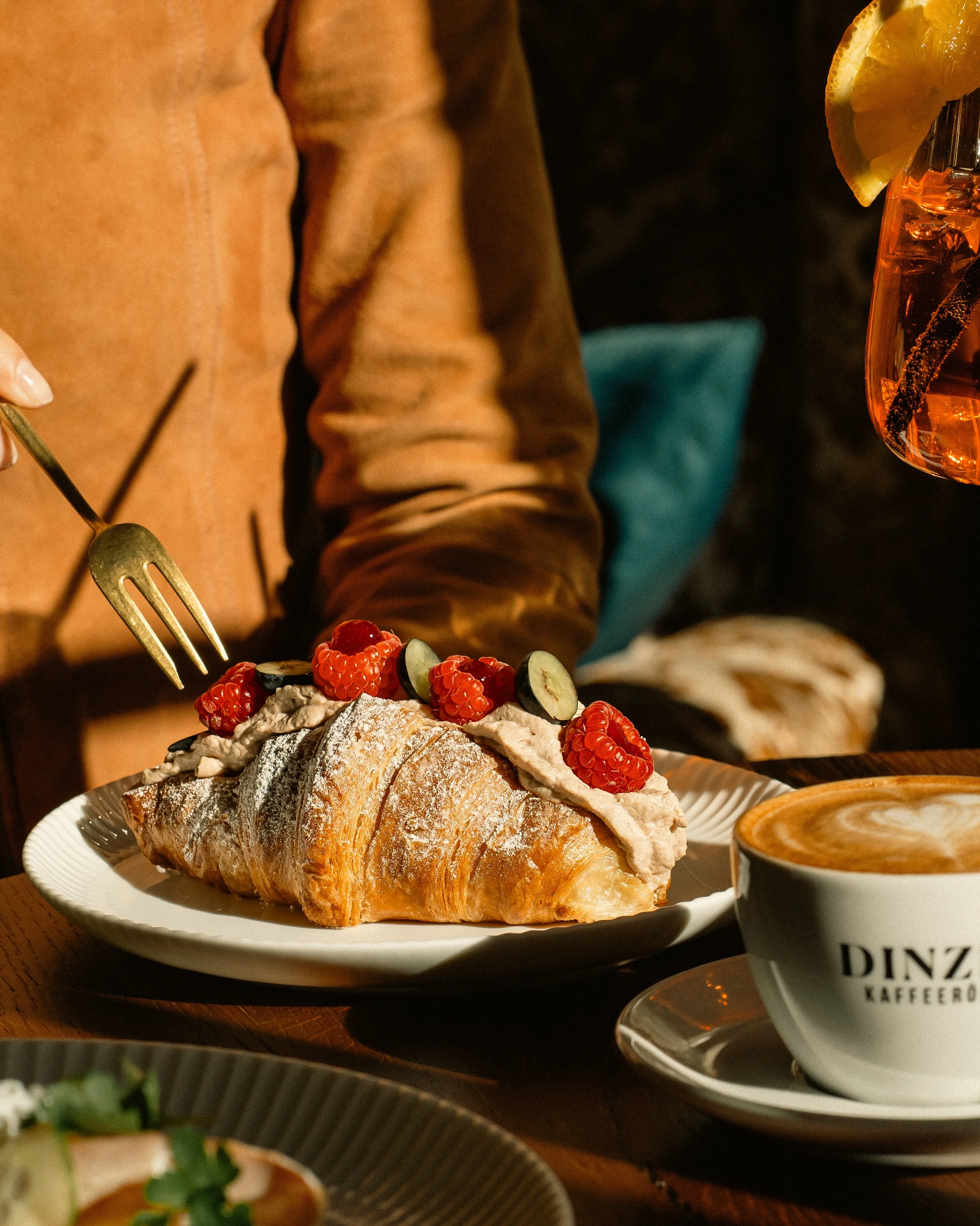 Person enjoying a croissant with cream and raspberries, a lemon-infused drink, and a cup of coffee on a wooden table.