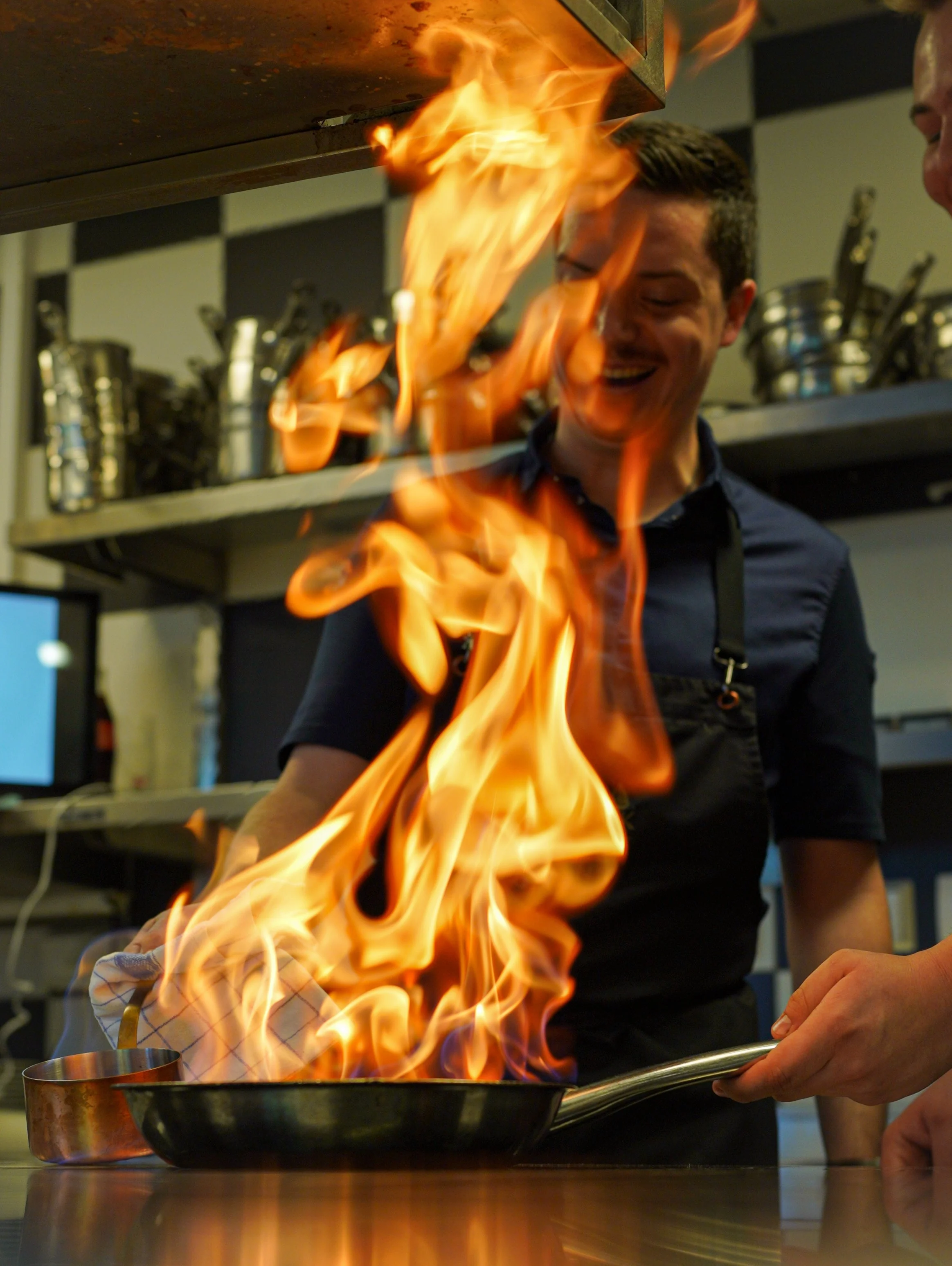 Chef holding a skillet with flames in a professional kitchen.
