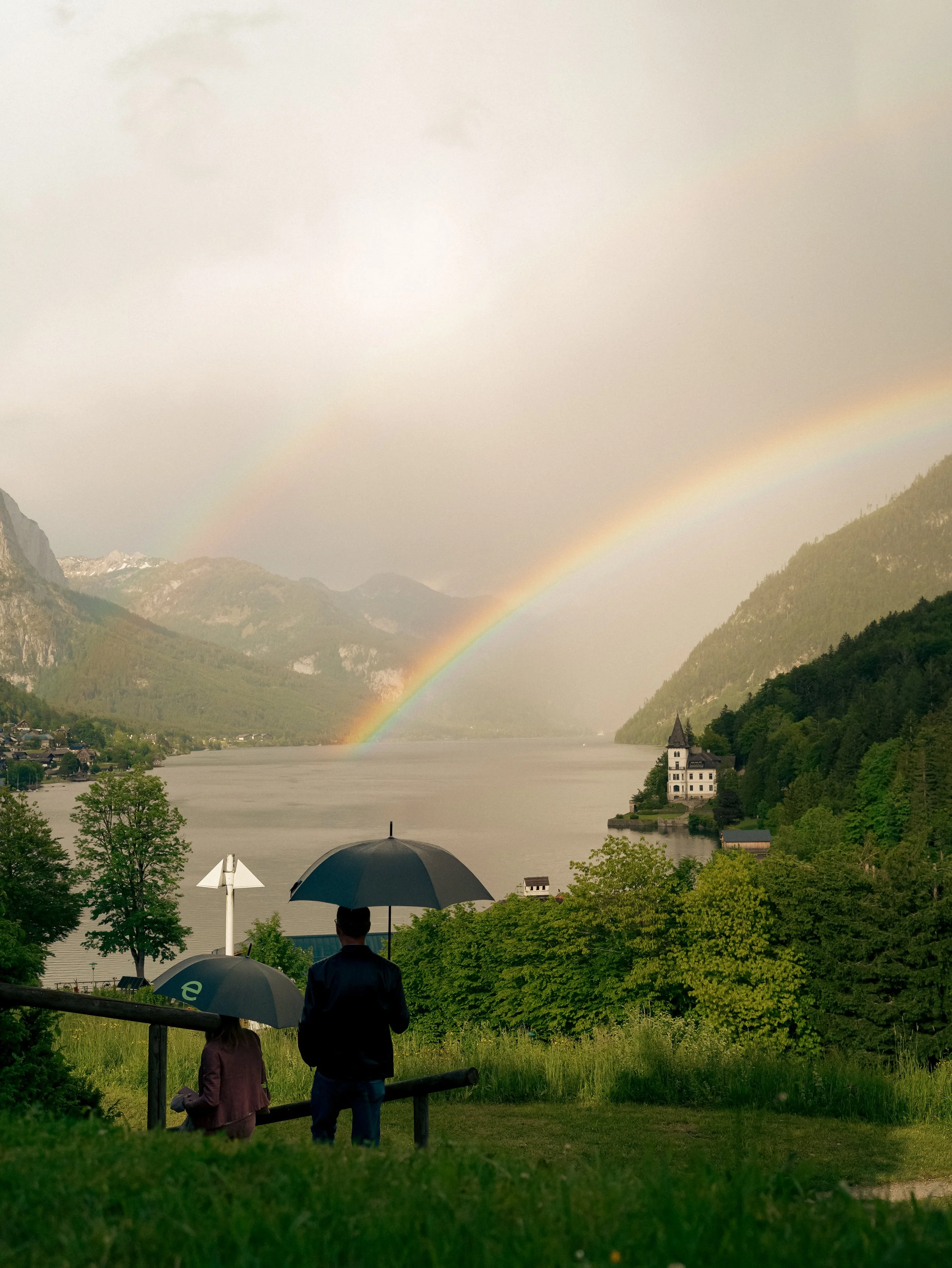 Two people with umbrellas overlooking a serene lake with a rainbow and distant mountains.
