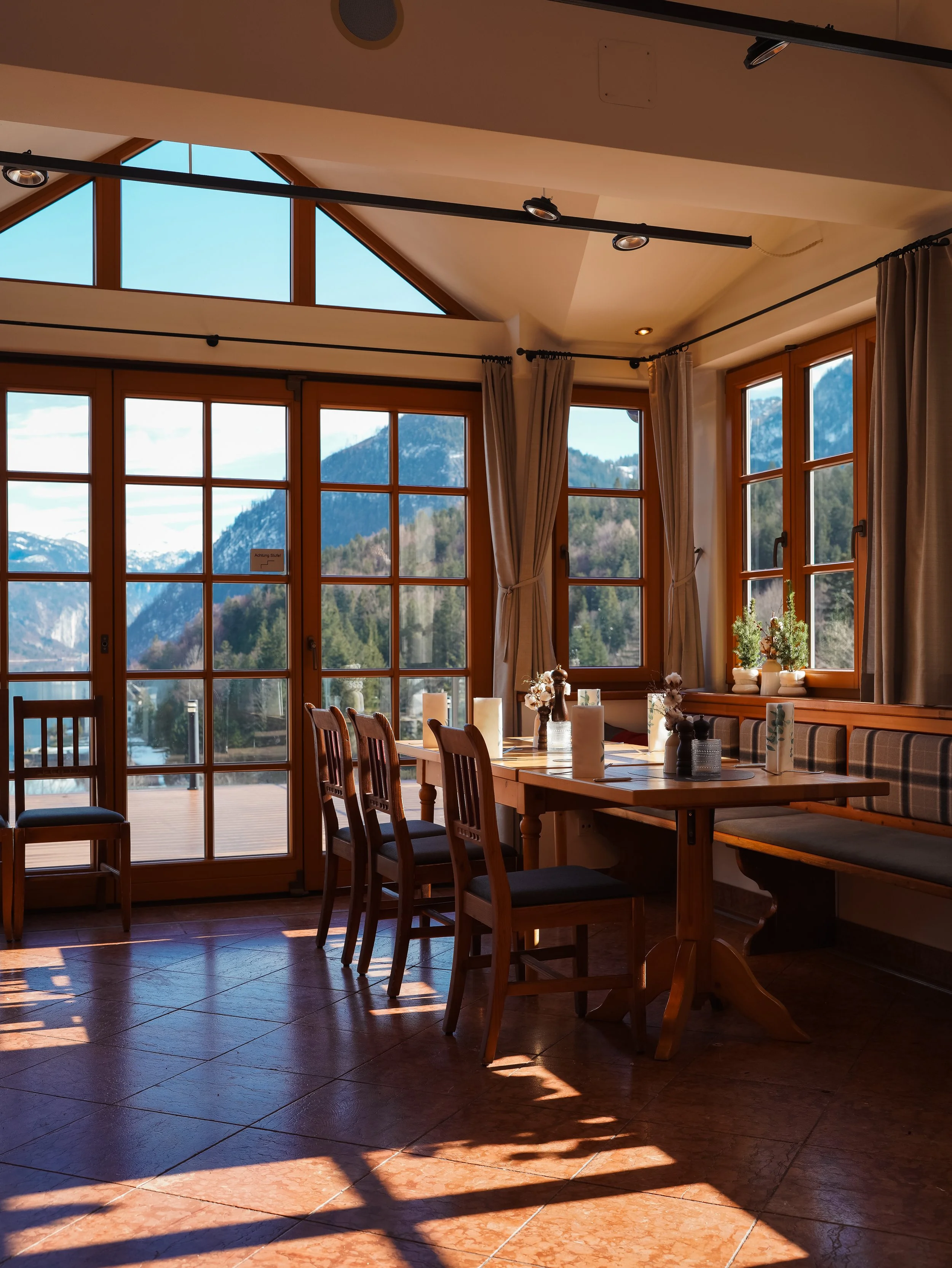 Cozy dining area with wooden table and chairs, large windows offering view of mountains, indoor plants and curtains, warm natural lighting.