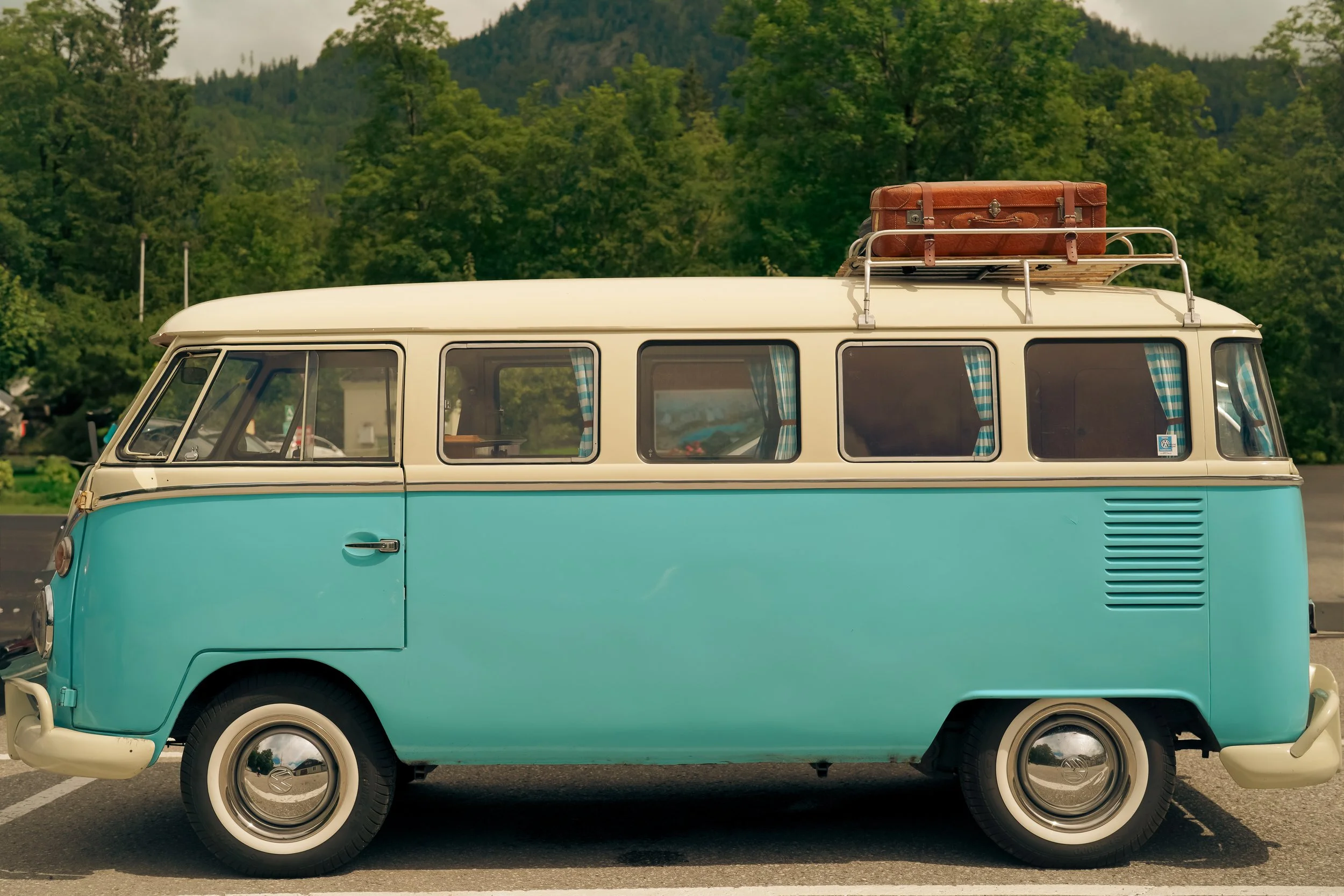 Vintage blue and cream van with a luggage rack and suitcase, parked in a scenic outdoor setting with trees and mountains.