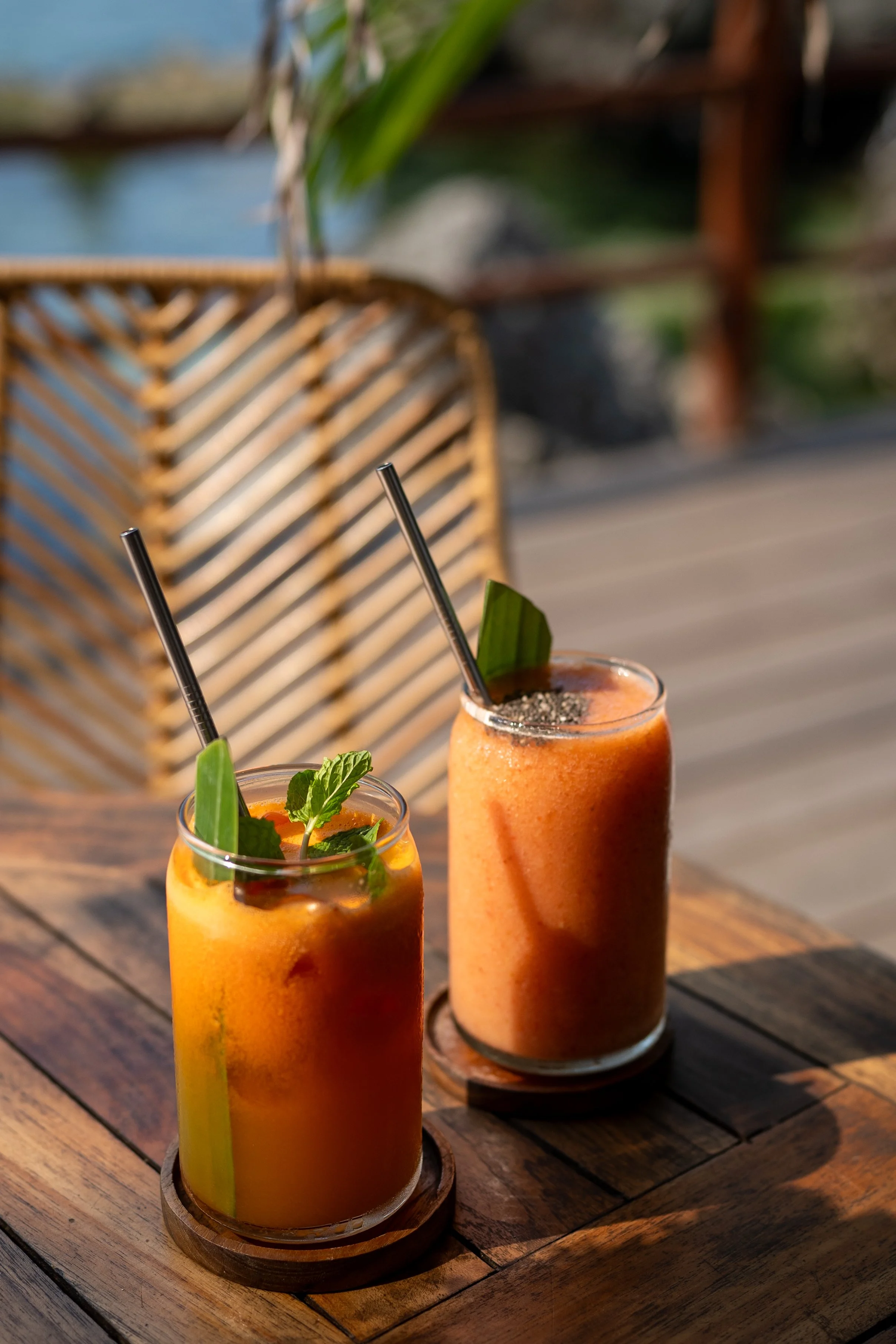 Two orange smoothies with mint garnish in glass cups on a wooden table, featuring metal straws and a wicker chair in the background.