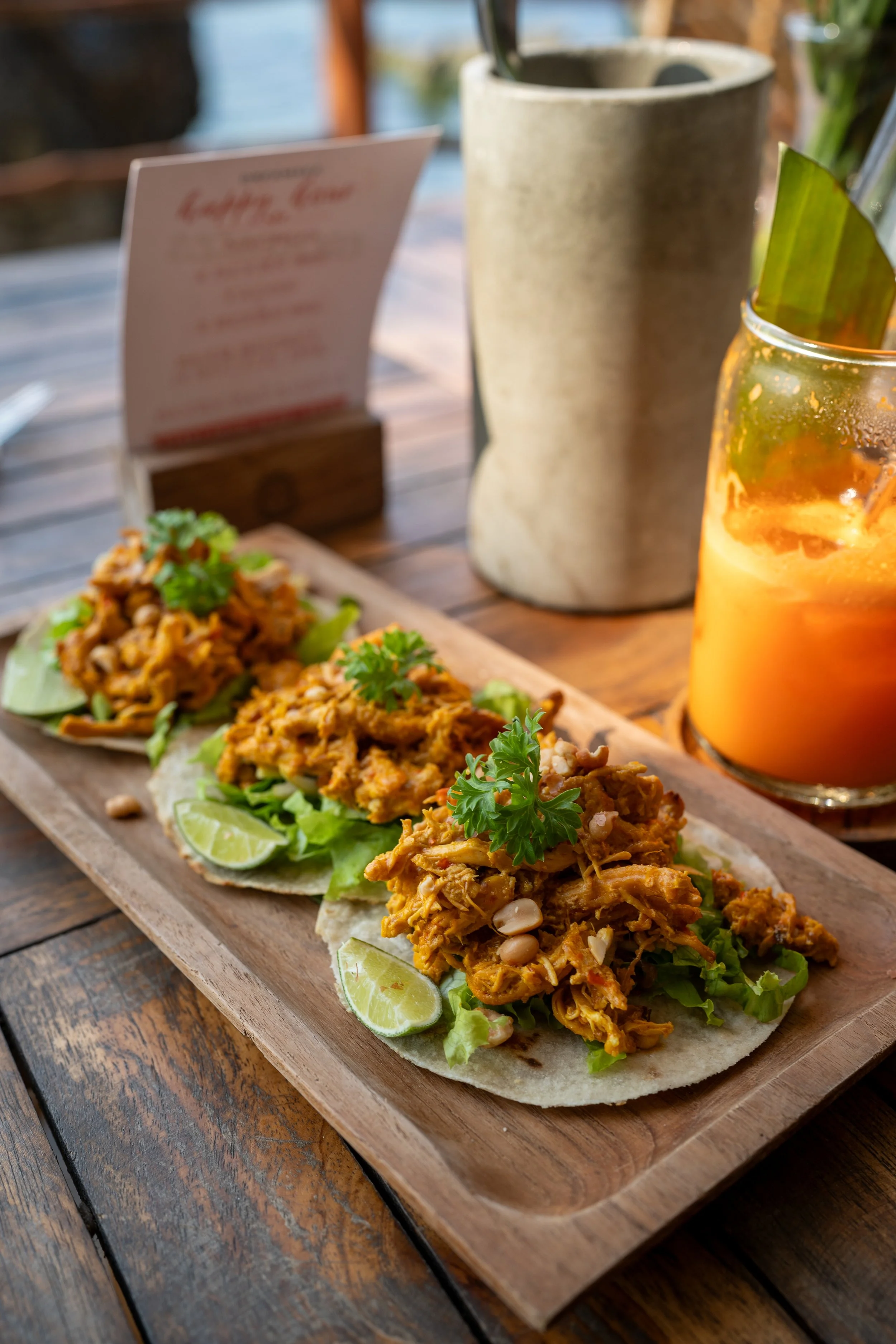 Three chicken tacos with lime slices and garnish on a wooden tray, accompanied by a glass of orange drink on a wooden table.