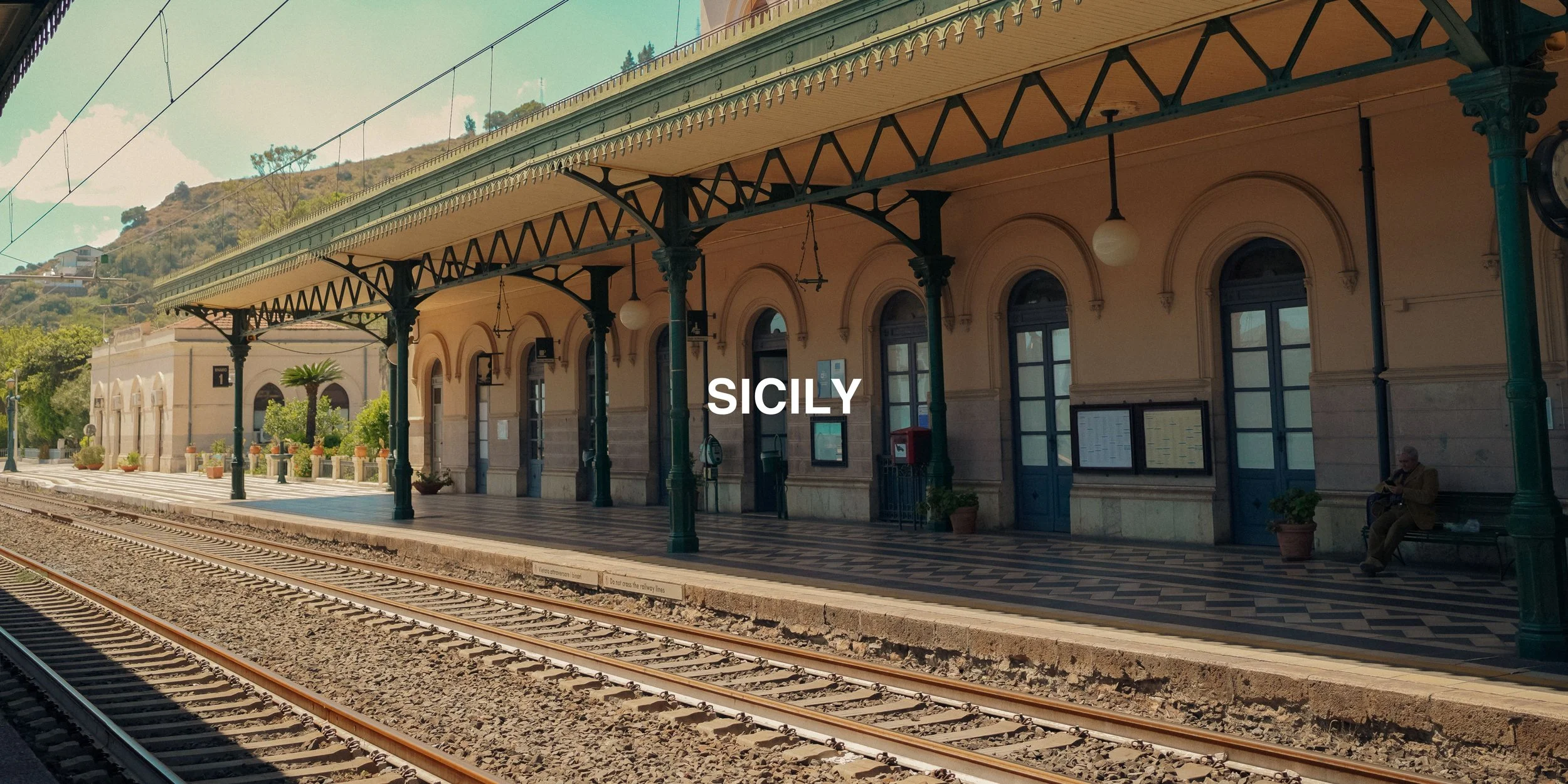 Train station in Sicily with vintage architecture and green columns, tracks in the foreground, hillside in the background.