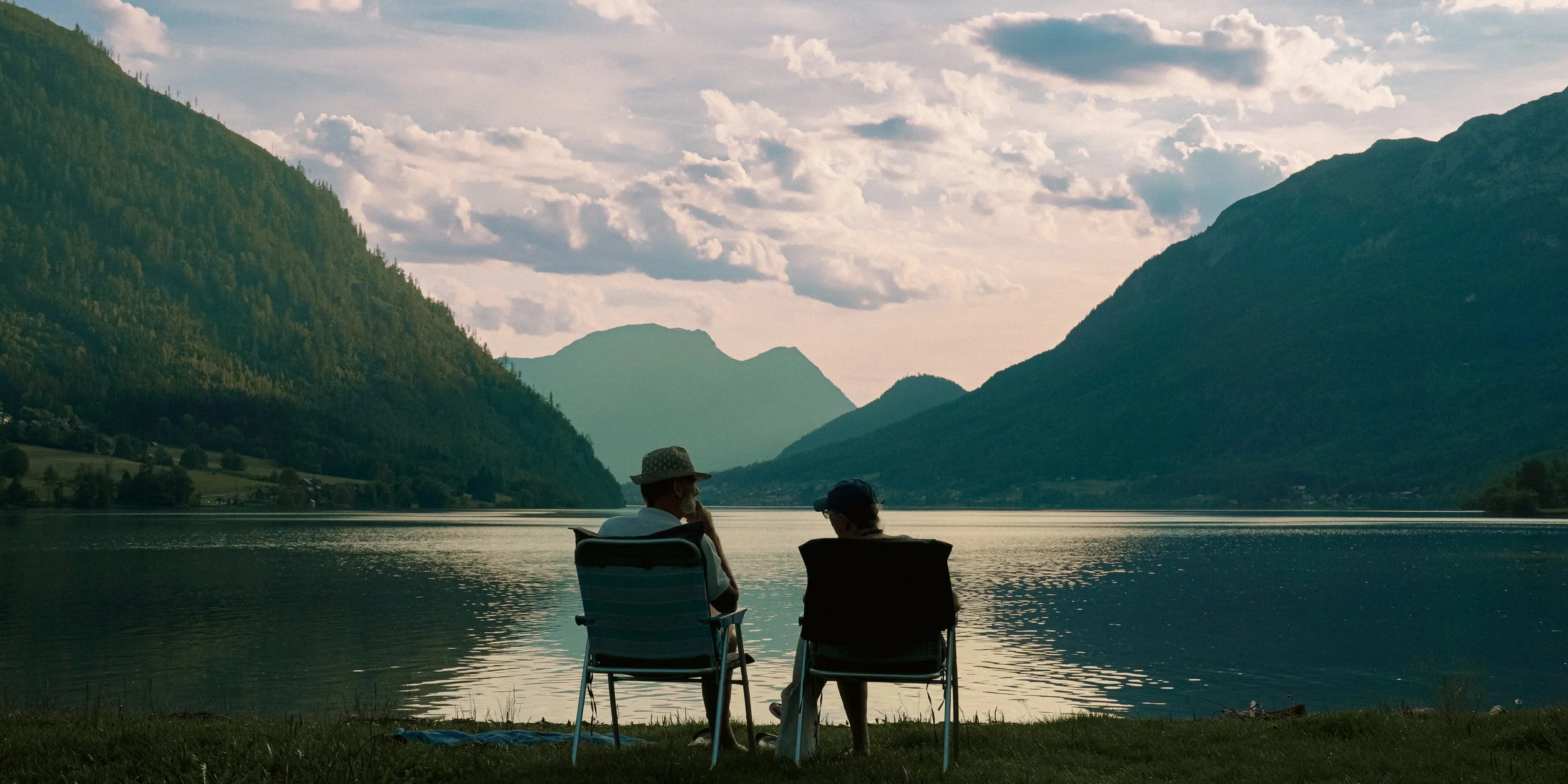 Two people sitting on chairs by a lake, surrounded by mountains, under a cloudy sky at sunset.