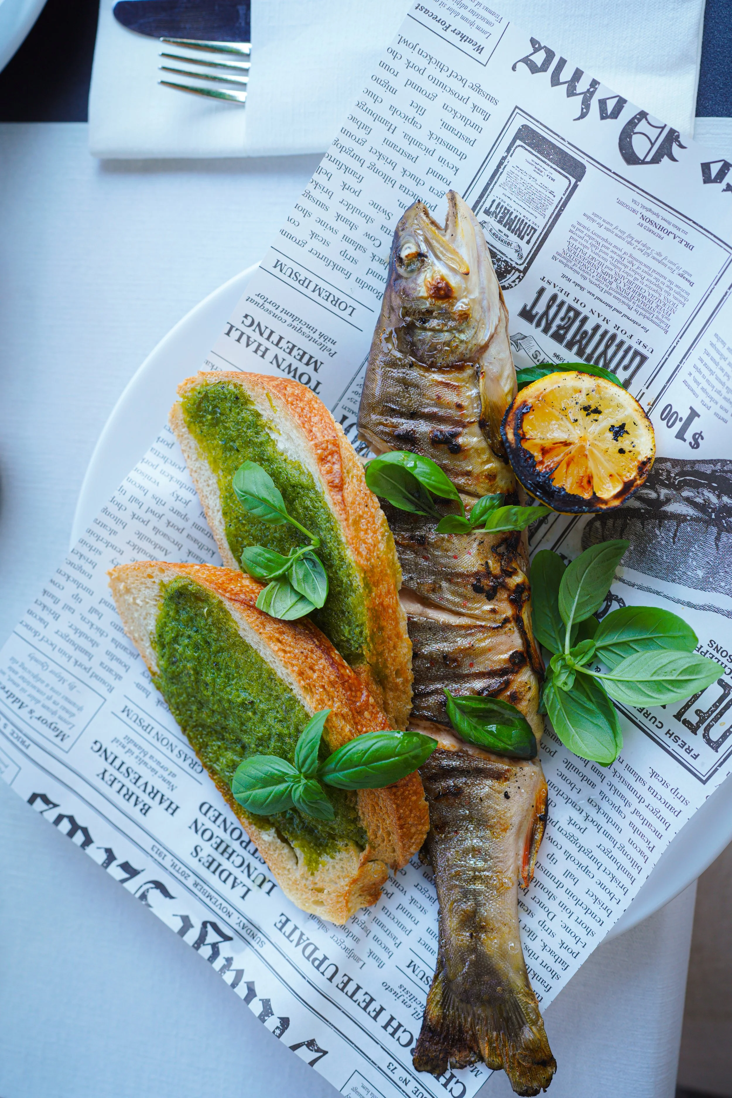 Grilled fish with lemon, pesto toast, and basil on newspaper-lined plate.