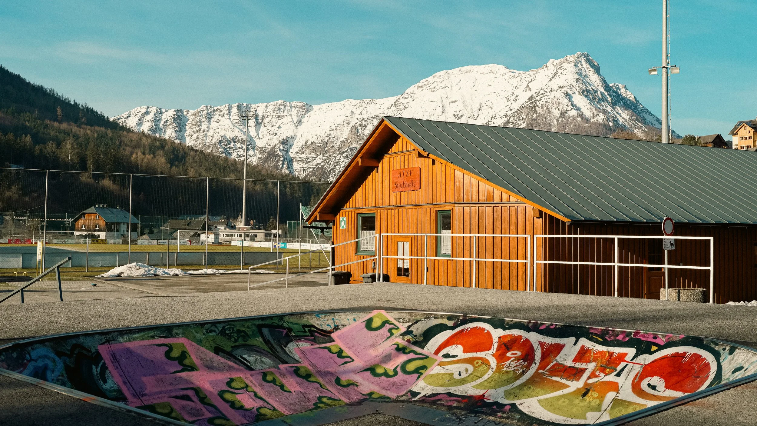 A wooden building with a gable roof stands near a skatepark with colorful graffiti. Snow-capped mountains are visible in the background under a clear blue sky.