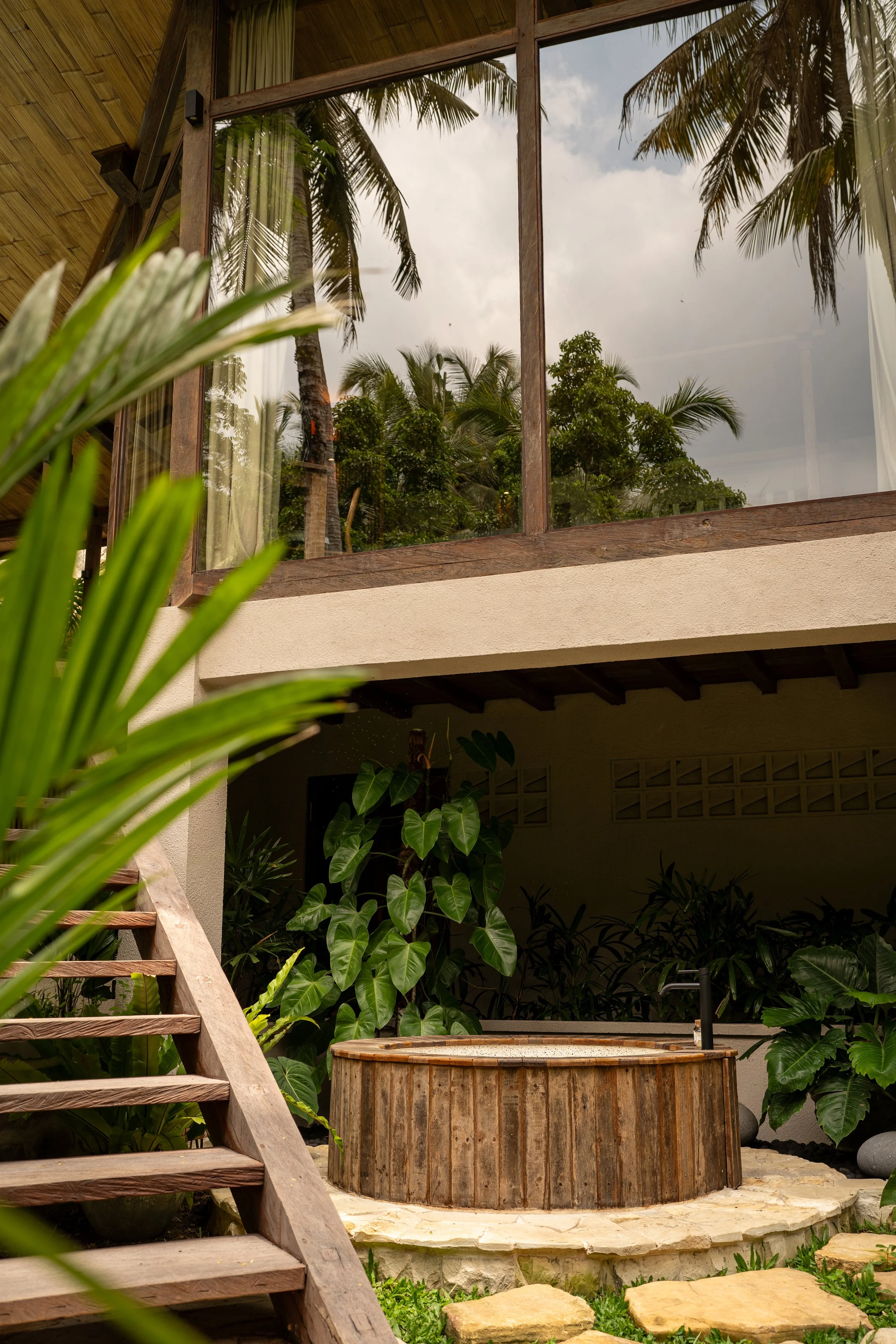 Outdoor spa area with rustic wooden hot tub surrounded by lush greenery and palm trees, reflecting in large glass windows of a wooden house.