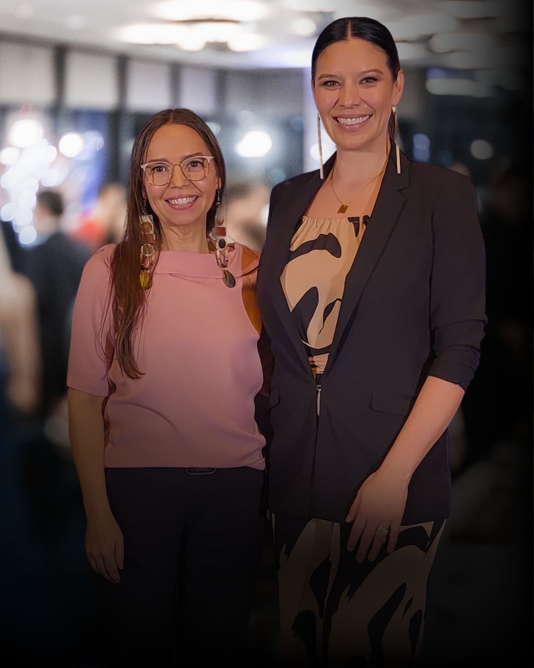 Two women smiling at an event, one wearing a pink top and glasses, the other in a black blazer with a patterned dress, in a dimly lit setting with blurred background