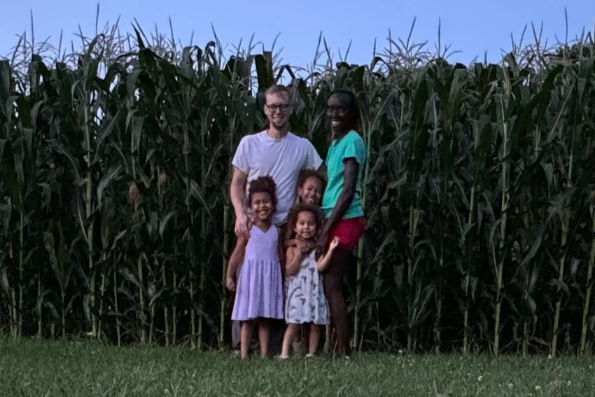A family of five stands in front of tall corn plants outdoors during dusk. The family includes two adults and three children, all smiling and dressed casually.