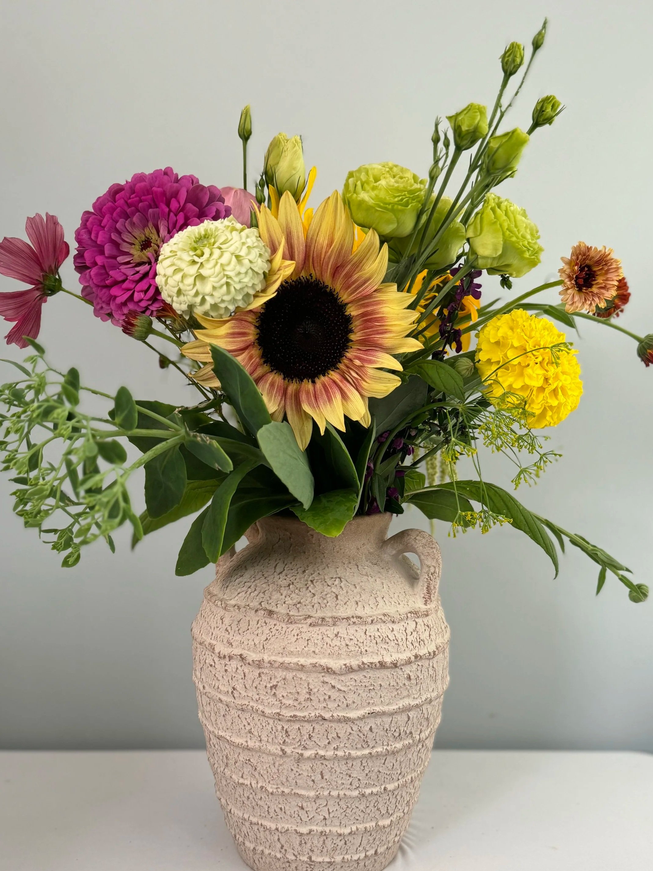 Colorful mixed flower bouquet in a textured beige ceramic vase placed on a white surface against a plain light background.