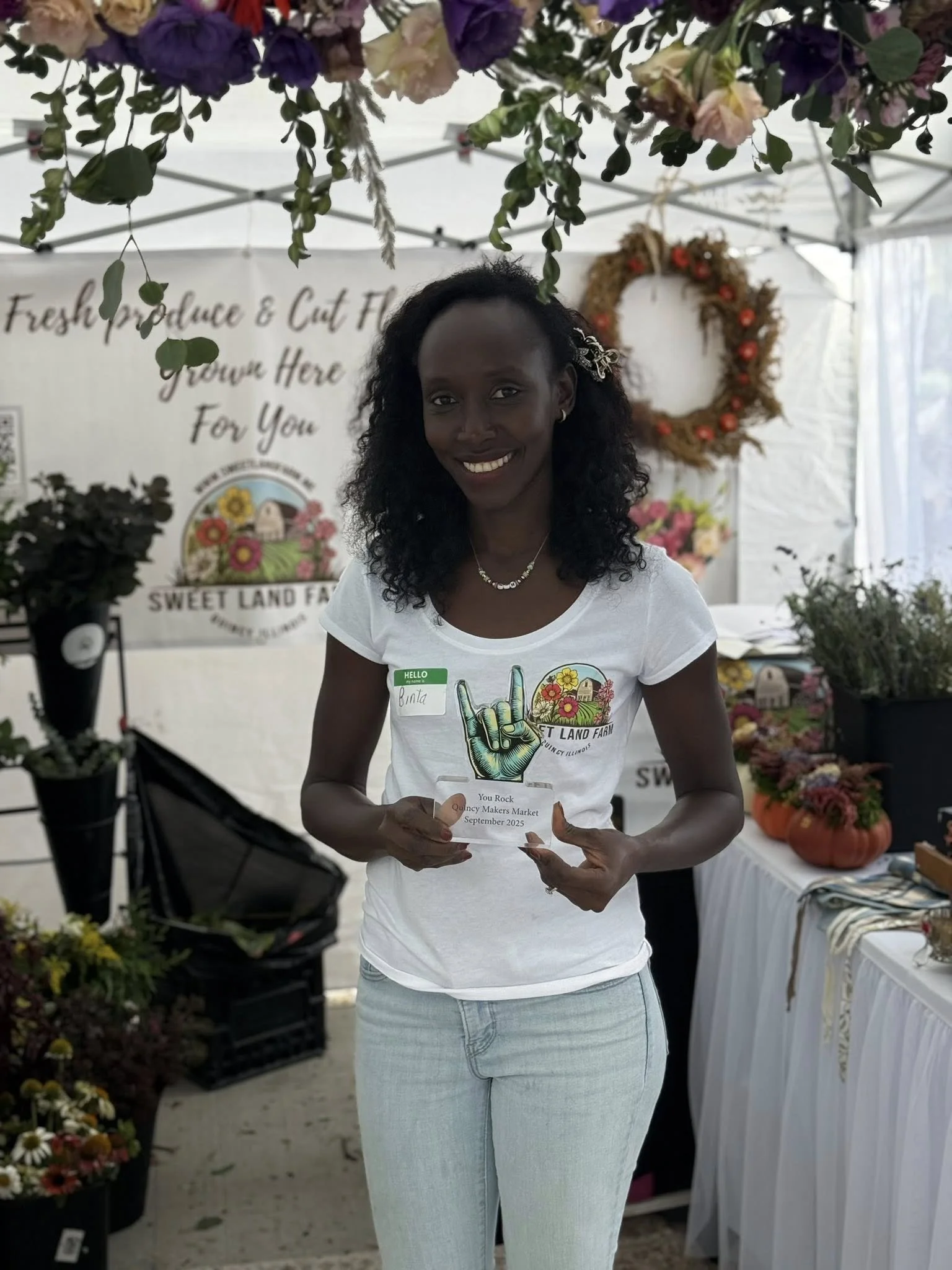 Woman holding a clear plastic award in front of a flower stand at an outdoor market. She is smiling, wearing a white t-shirt with a graphic of a hand making a rock sign, and light blue jeans.