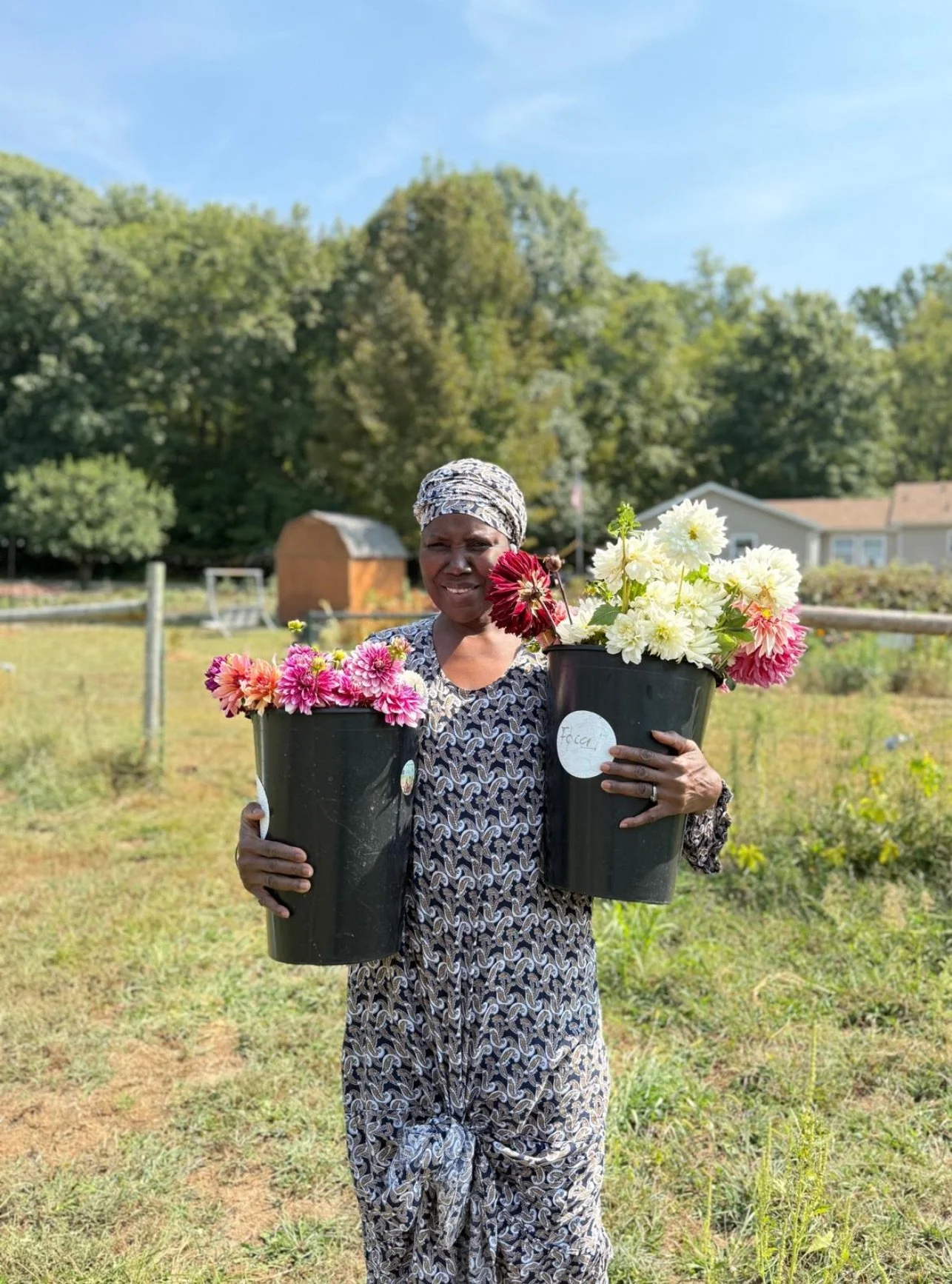 A woman standing outdoors in a garden holding two large black pots filled with colorful dahlias, smiling at the camera.