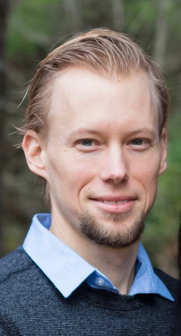Close-up of a young man with light skin, light brown hair, and a goatee, wearing a light blue collared shirt and a dark sweater, outdoors with blurred trees in the background.