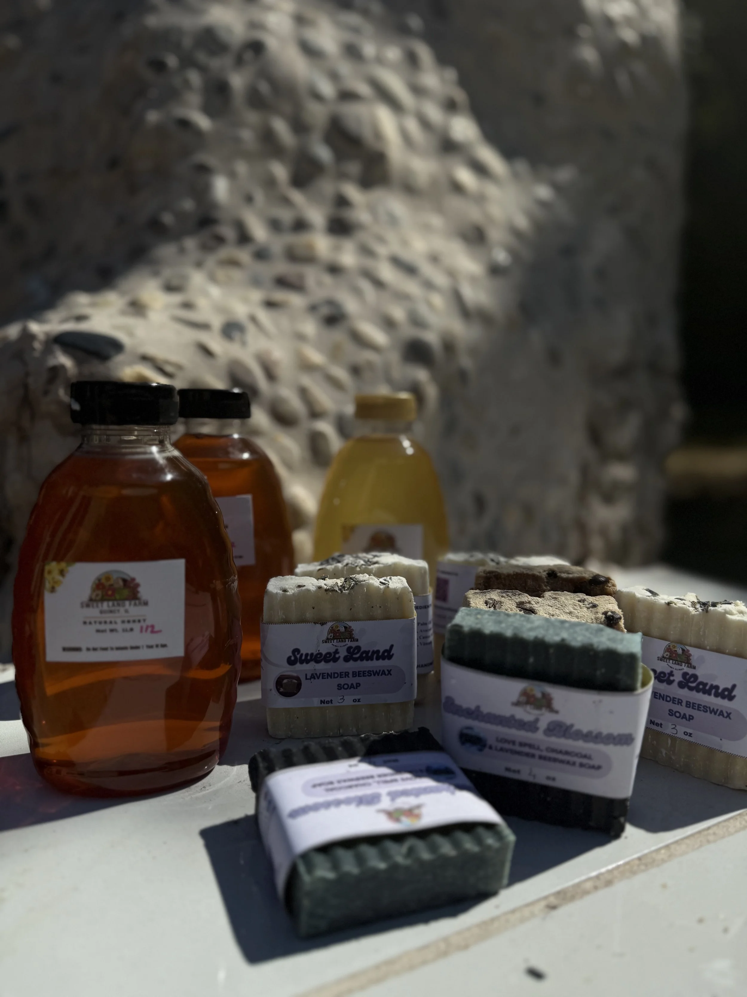 Assorted handmade soaps and honey jars displayed outdoors on a white surface with a stone sculpture of an animal in the background.