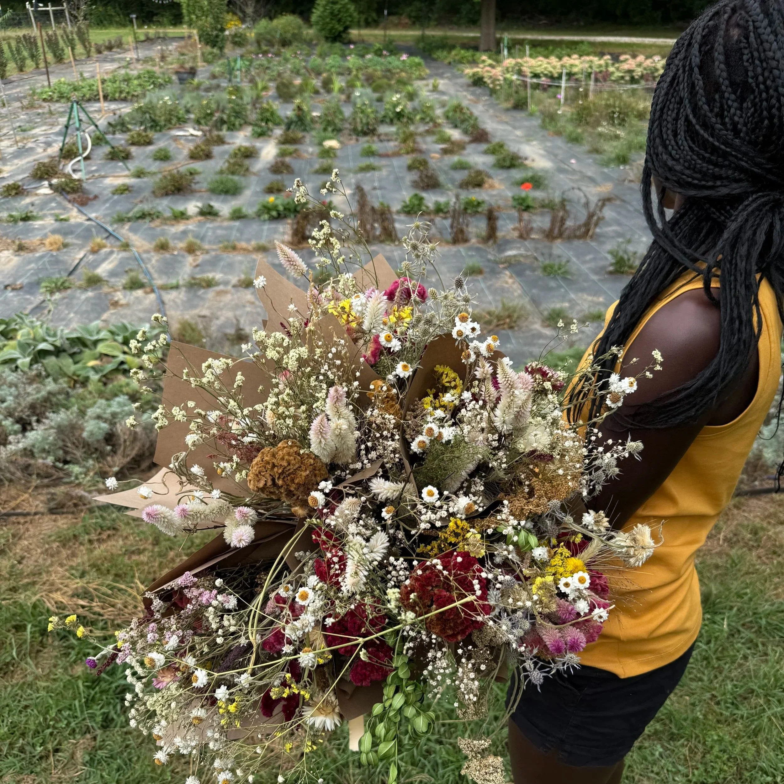 A woman holding a large bouquet of mixed flowers, standing in a garden with planted rows in the background.