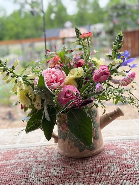 A bouquet of pink, purple, white, and yellow flowers in a small watering can on a patterned tablecloth.