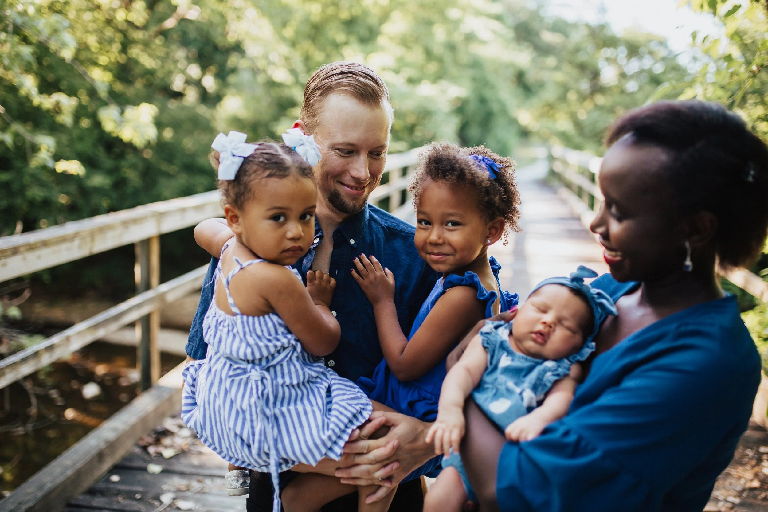A family of five, including a father, mother, and three young children, stands on a wooden bridge in a lush green forest. The father and mother are holding their two daughters and a sleeping baby girl, all dressed in blue outfits, celebrating a family moment outdoors.