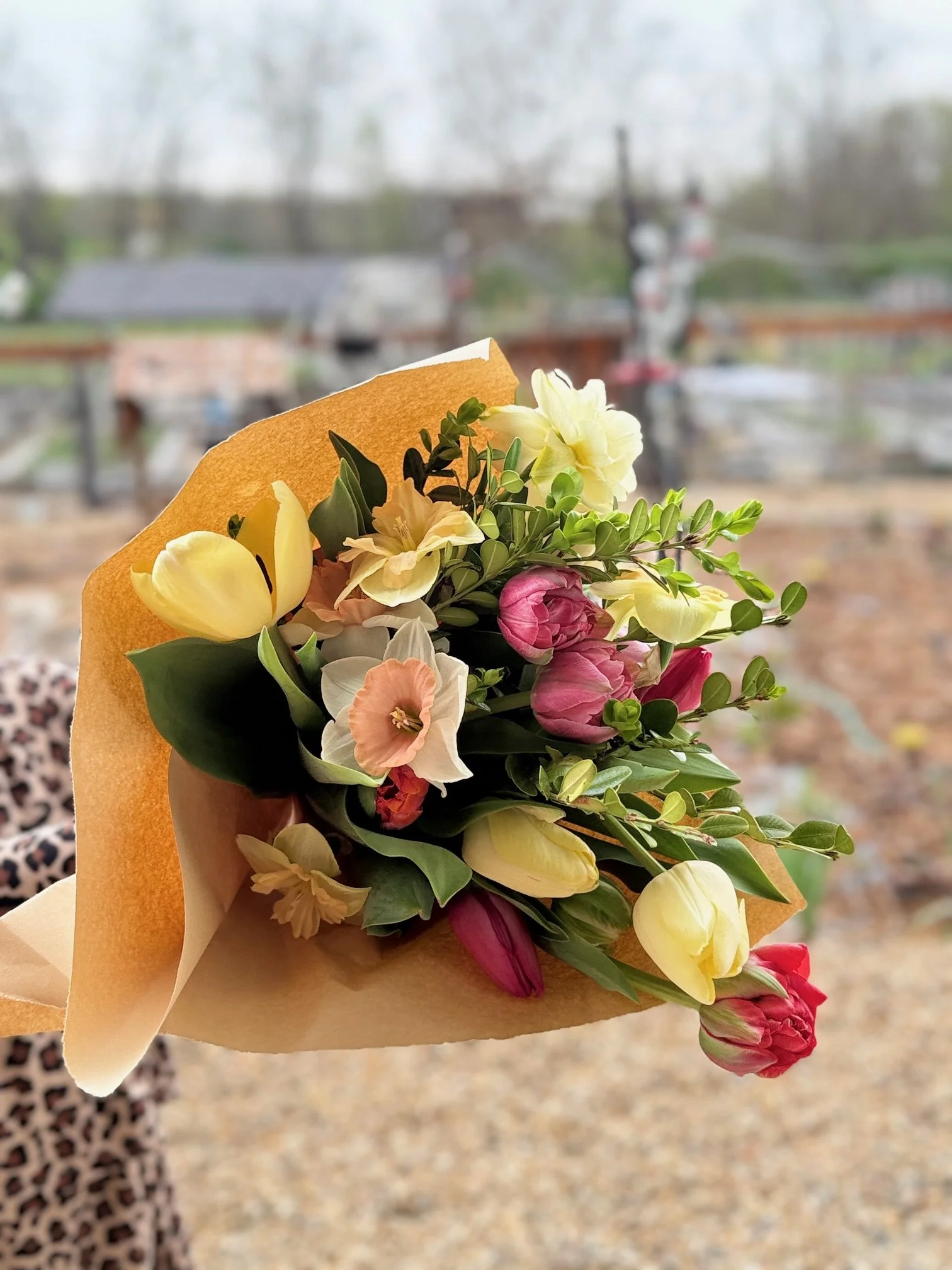 A bouquet of colorful flowers, including tulips and other blossoms, wrapped in yellowish paper, held outdoors with a blurred background of trees and a train crossing.
