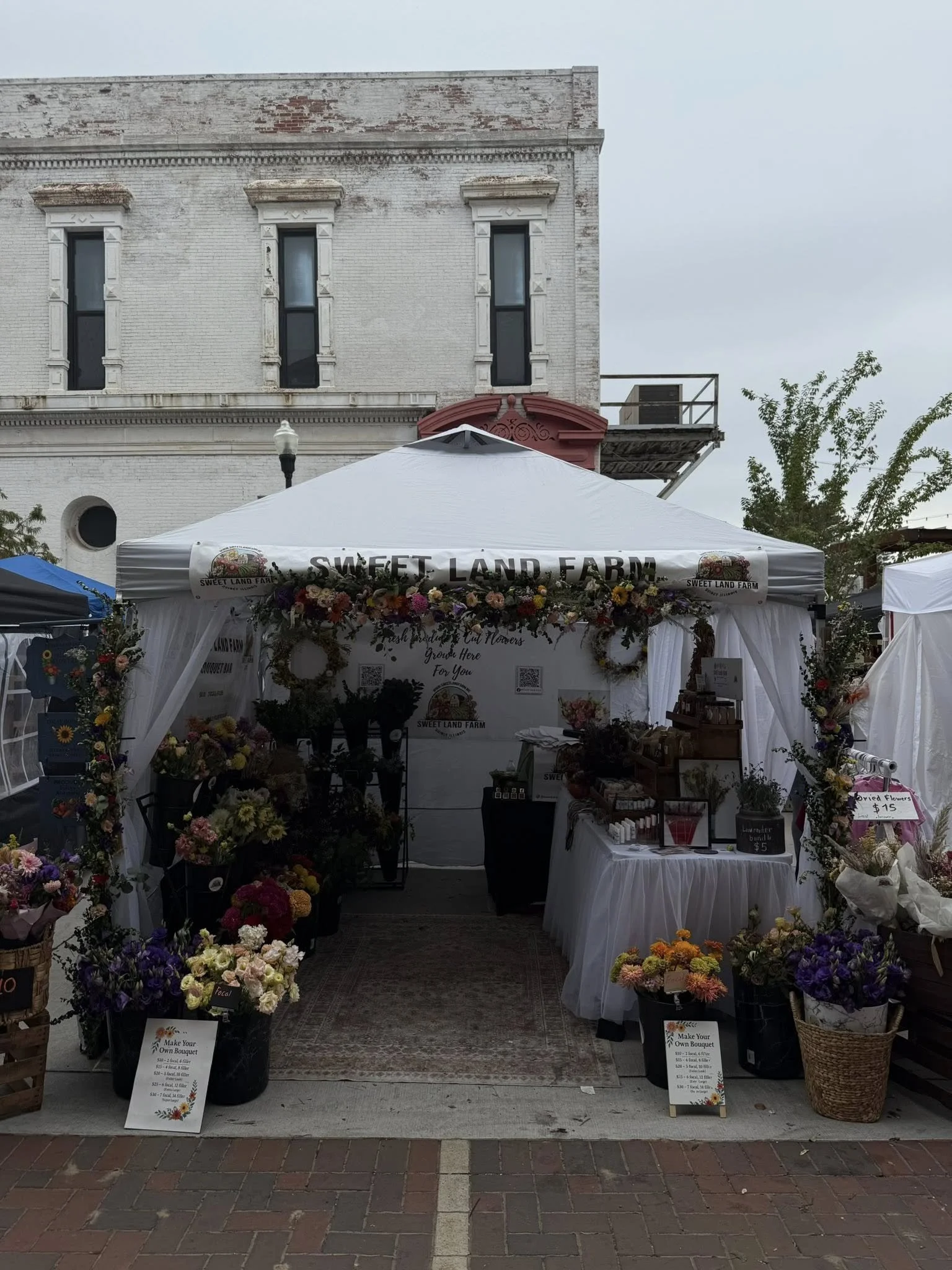 A flower stall at an outdoor market with a white canopy that reads 'Sweet Land Farm.' The stall is decorated with colorful flowers and has various small bouquets and arrangements for sale. There are signs indicating prices for dried flowers and making your own bouquet. The background includes a brick sidewalk and a white brick building with tall windows.