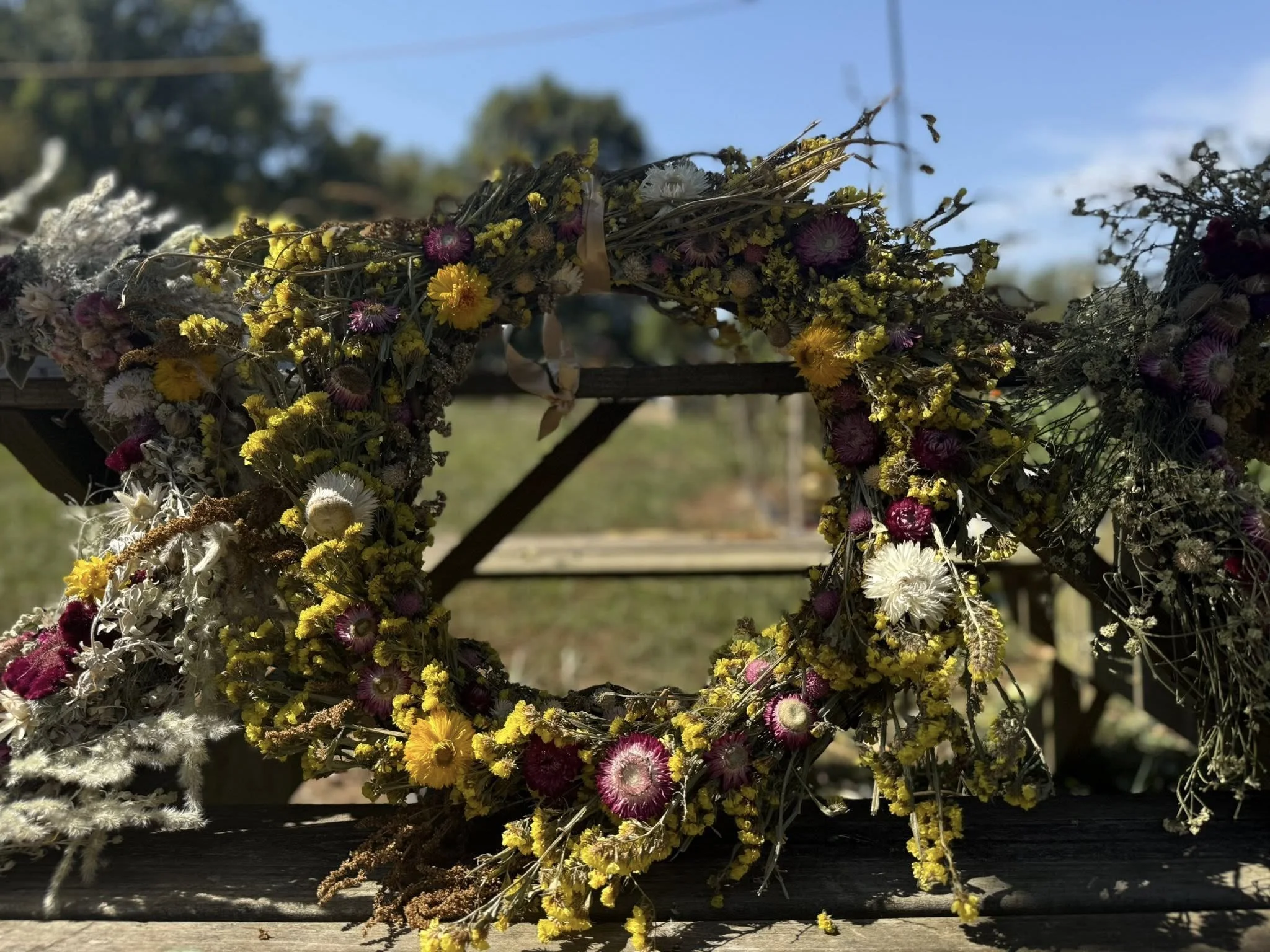 A floral wreath made of yellow, purple, white, and pink dried flowers on a rustic wooden surface outdoors with a blurred background of trees and blue sky.