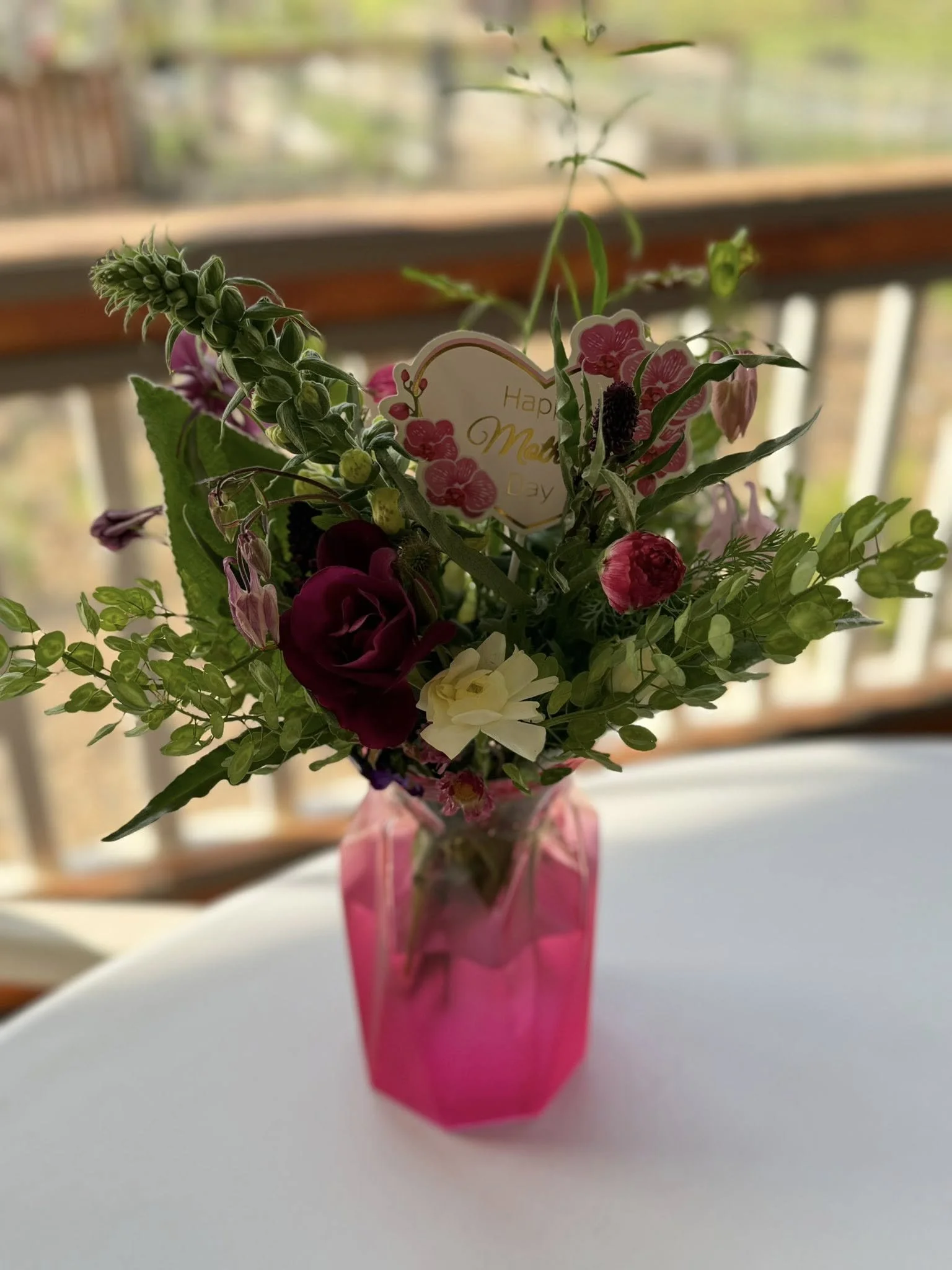 Fresh flower bouquet in a bright pink vase, with a small card that says "Happy Mother's Day" on a white table, with a blurred background of a balcony and outdoor scenery.