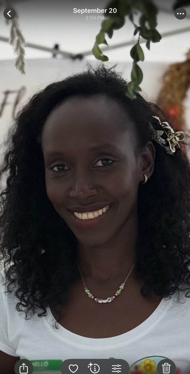 A smiling woman with dark curly hair wearing a necklace and a hair accessory, indoors with hanging plants and a decorated wall in the background.