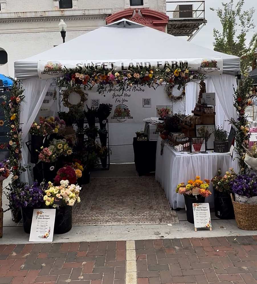 A flower stand called 'Sweet Land Farm' decorated with colorful flowers and floral arrangements at an outdoor market or fair.