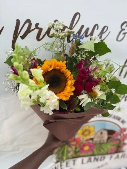 A colorful bouquet of flowers, including a sunflower, white, purple, and yellow blossoms, wrapped in brown paper.
