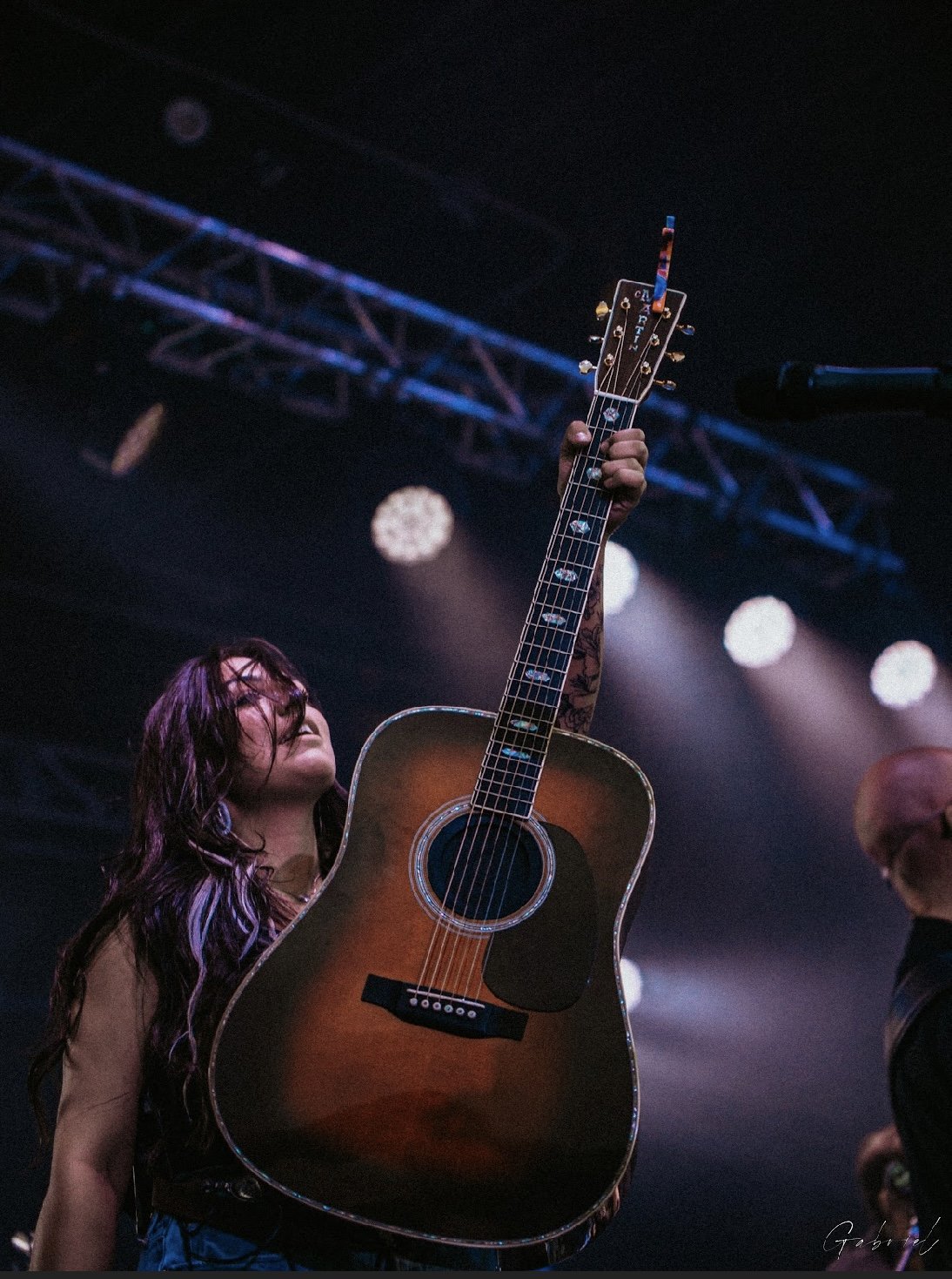 Booking Promo #10. Emily Raff holding an acoustic guitar on stage with stage lighting in the background.