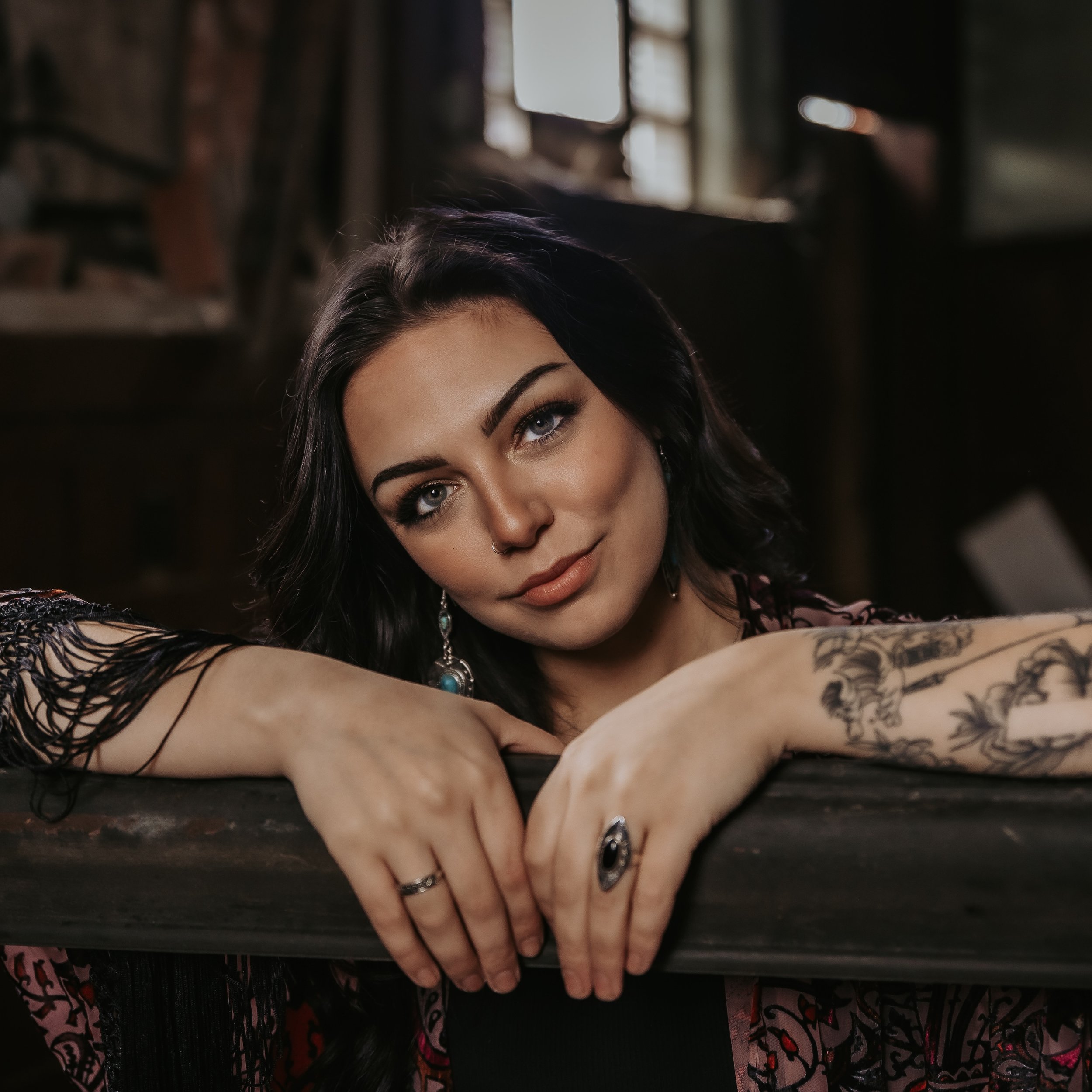 Booking Promo #6. Emily Raff with dark hair and tattoos leaning on a railing indoors.