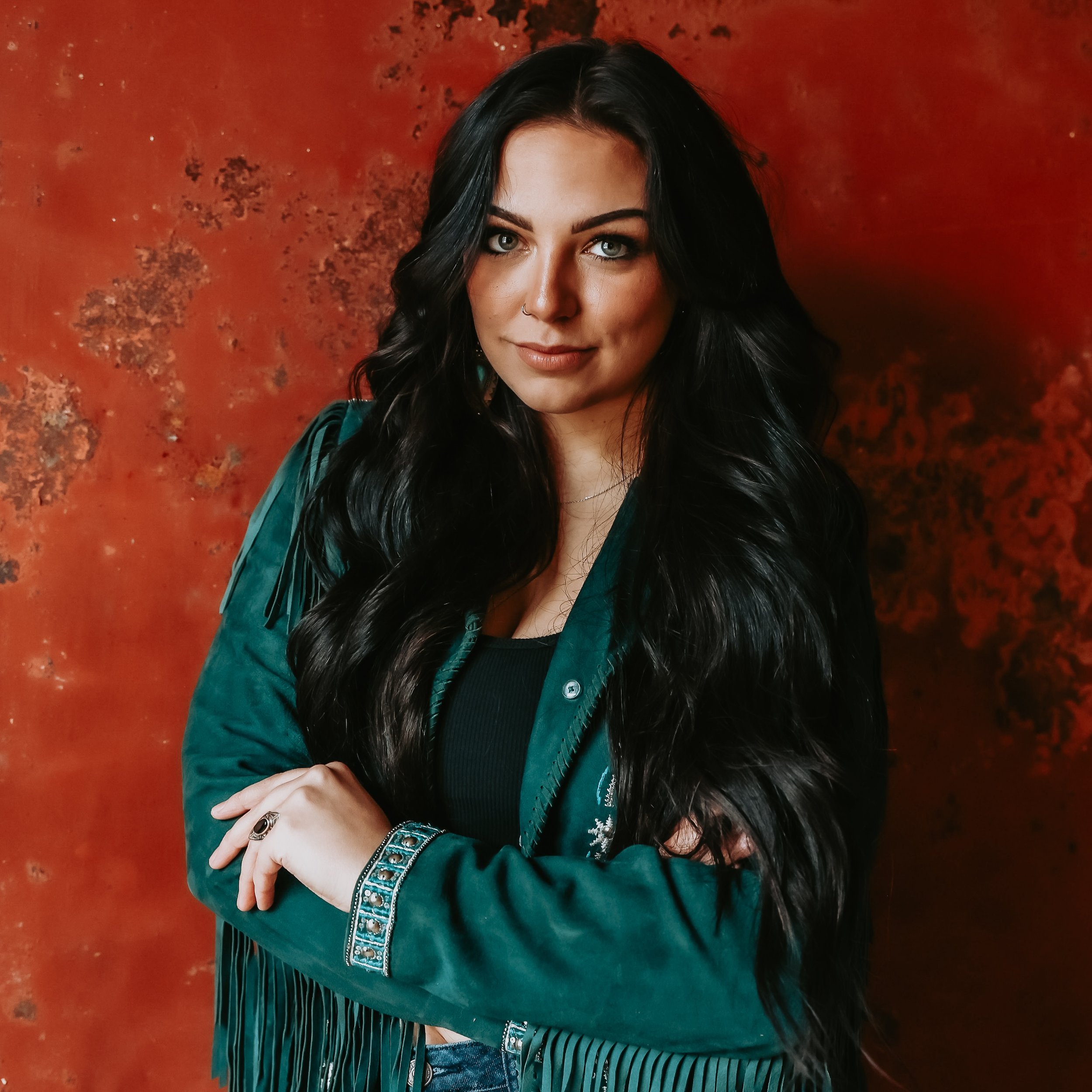 Booking Promo #4. Emily Raff with long dark hair in a green fringed jacket standing against a textured red wall.